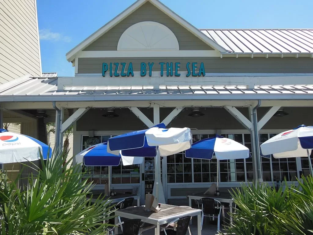 Front of a seafood restaurant called 'Pizza By The Sea' with outdoor seating and umbrellas, and a clear blue sky.