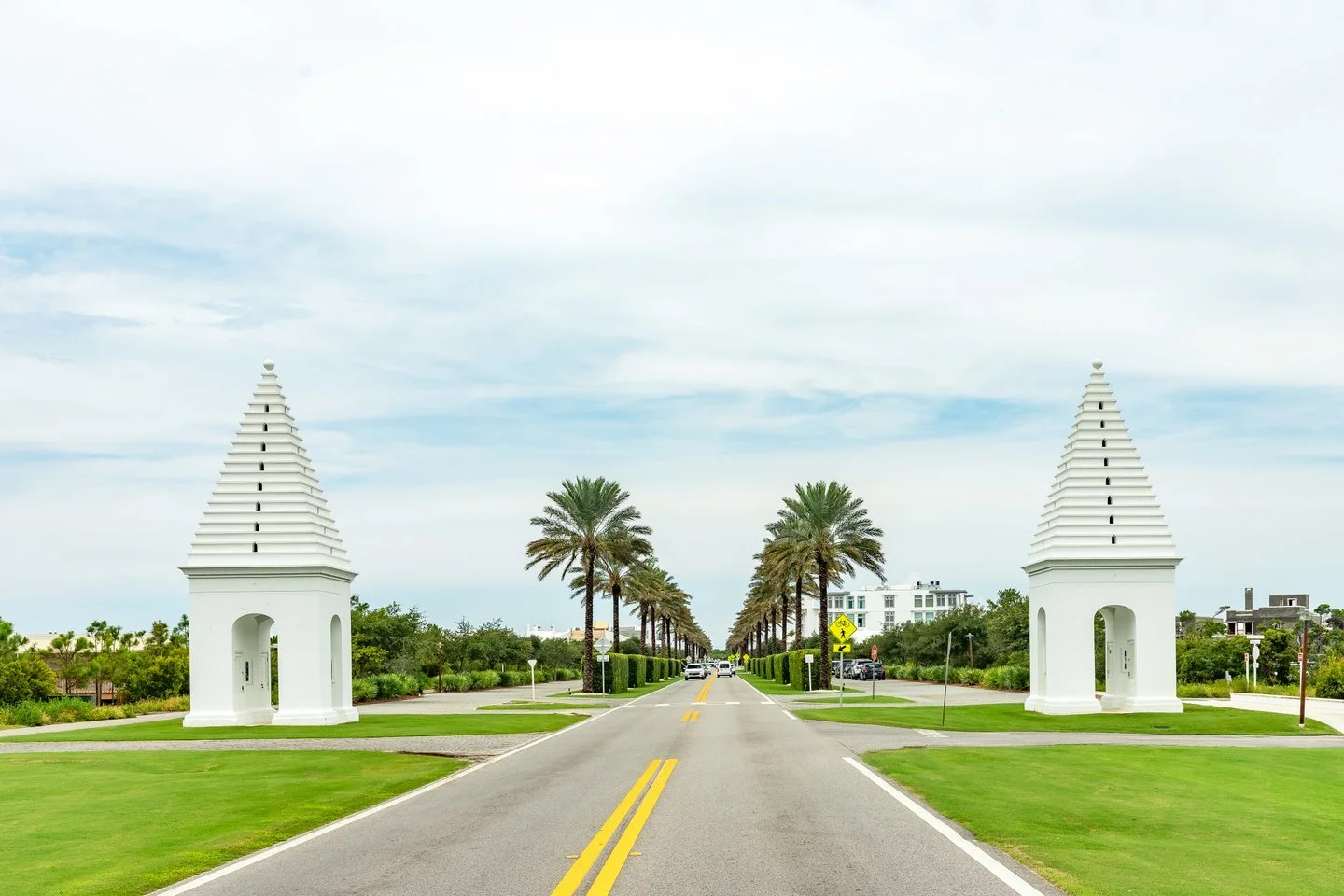 A symmetrical street lined with palm trees leading to white structures with tiered roofs, resembling gateways, on either side.