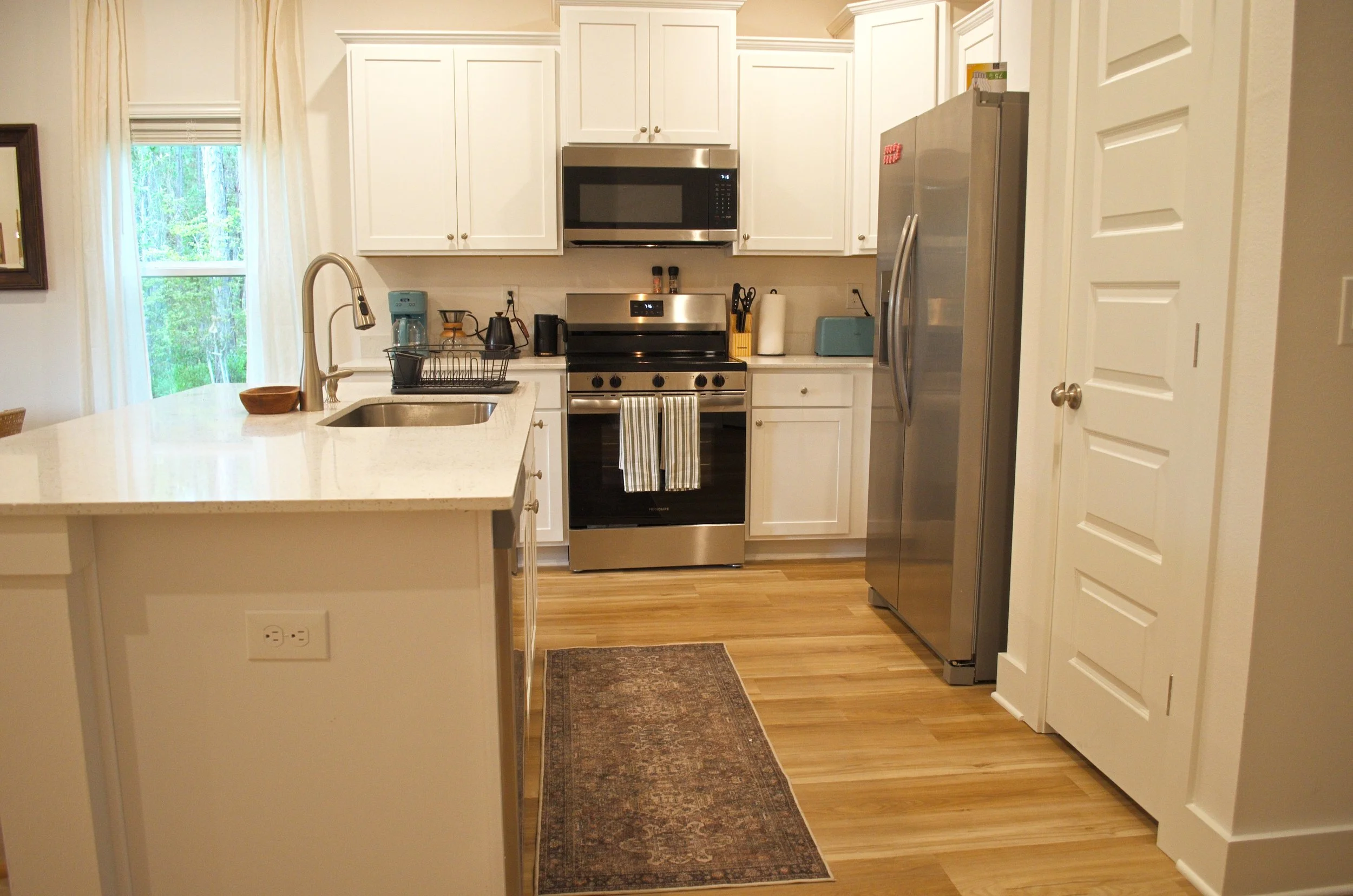 A kitchen with white cabinets, a stainless steel refrigerator, oven, microwave, a window with curtains, and various small appliances on the counter.