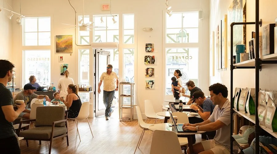People inside a bright, modern cafe with large windows, sitting at tables working on laptops, chatting, or enjoying coffee. The cafe has a minimalist decor with artwork on the walls and shelves with items for sale.