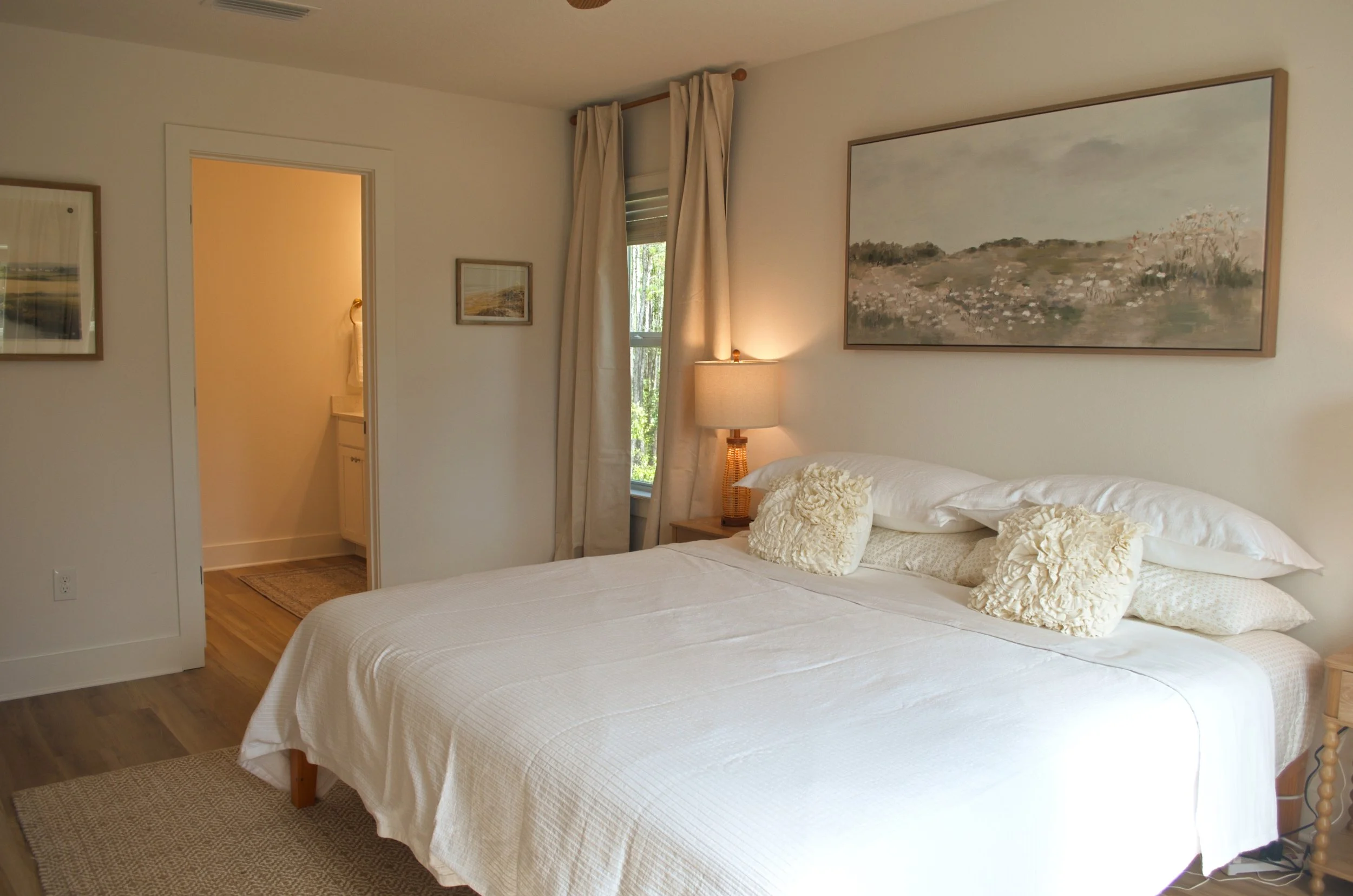 A cozy bedroom with a bed made with white bedding and cream pillows, a wooden nightstand with a lamp, and artwork on the walls. There's a window with cream curtains and a view of greenery outside.
