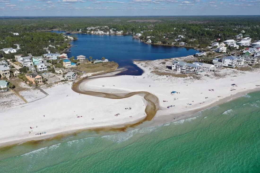 Aerial view of a beach with white sand, a creek running through the sand, and a residential area with houses near the shoreline. There is a lake behind the houses and the ocean in front.