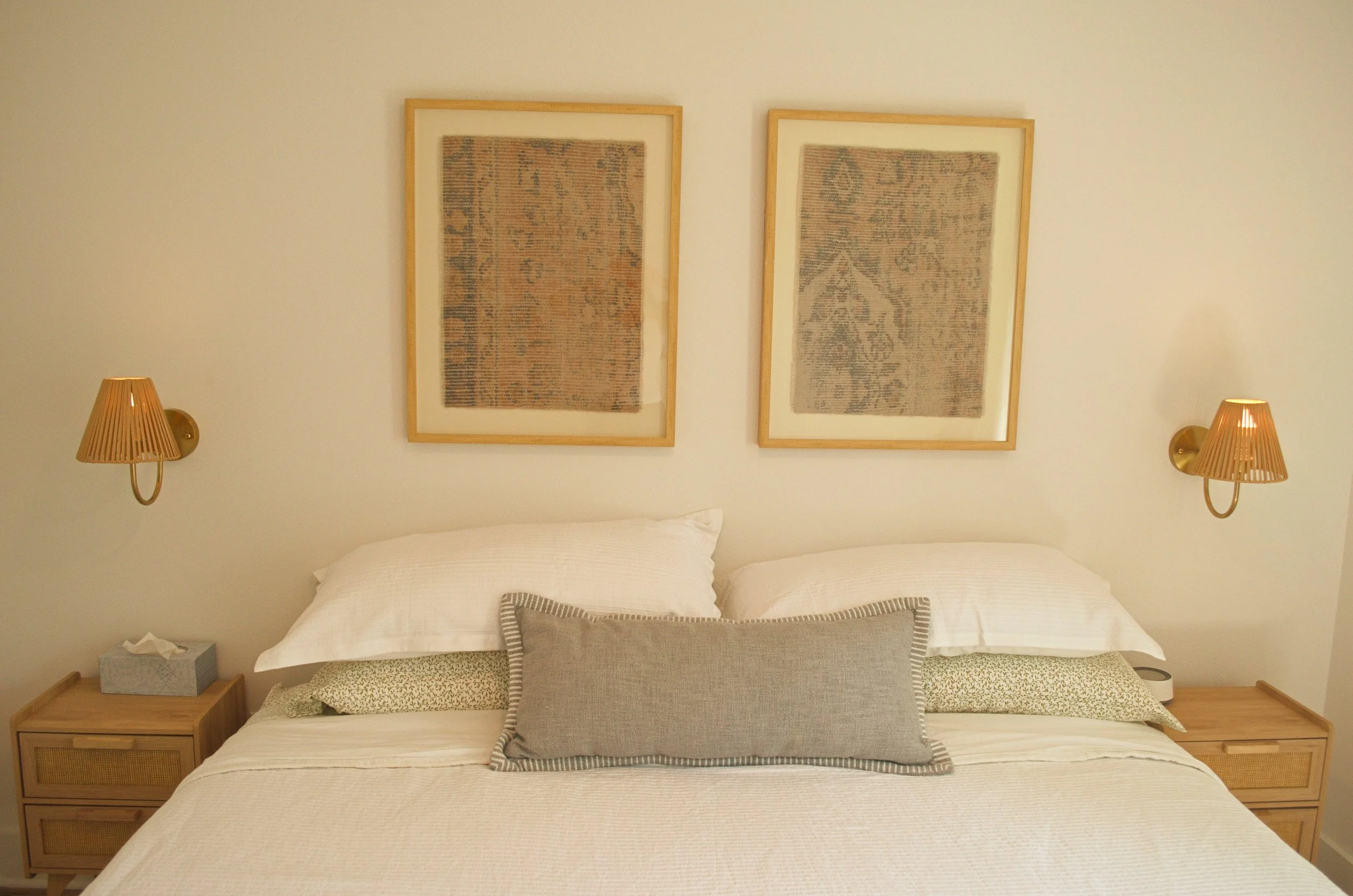 A neatly made bed with multiple pillows against a white wall, decorated with two framed textile art pieces and two wall-mounted lamps on either side.