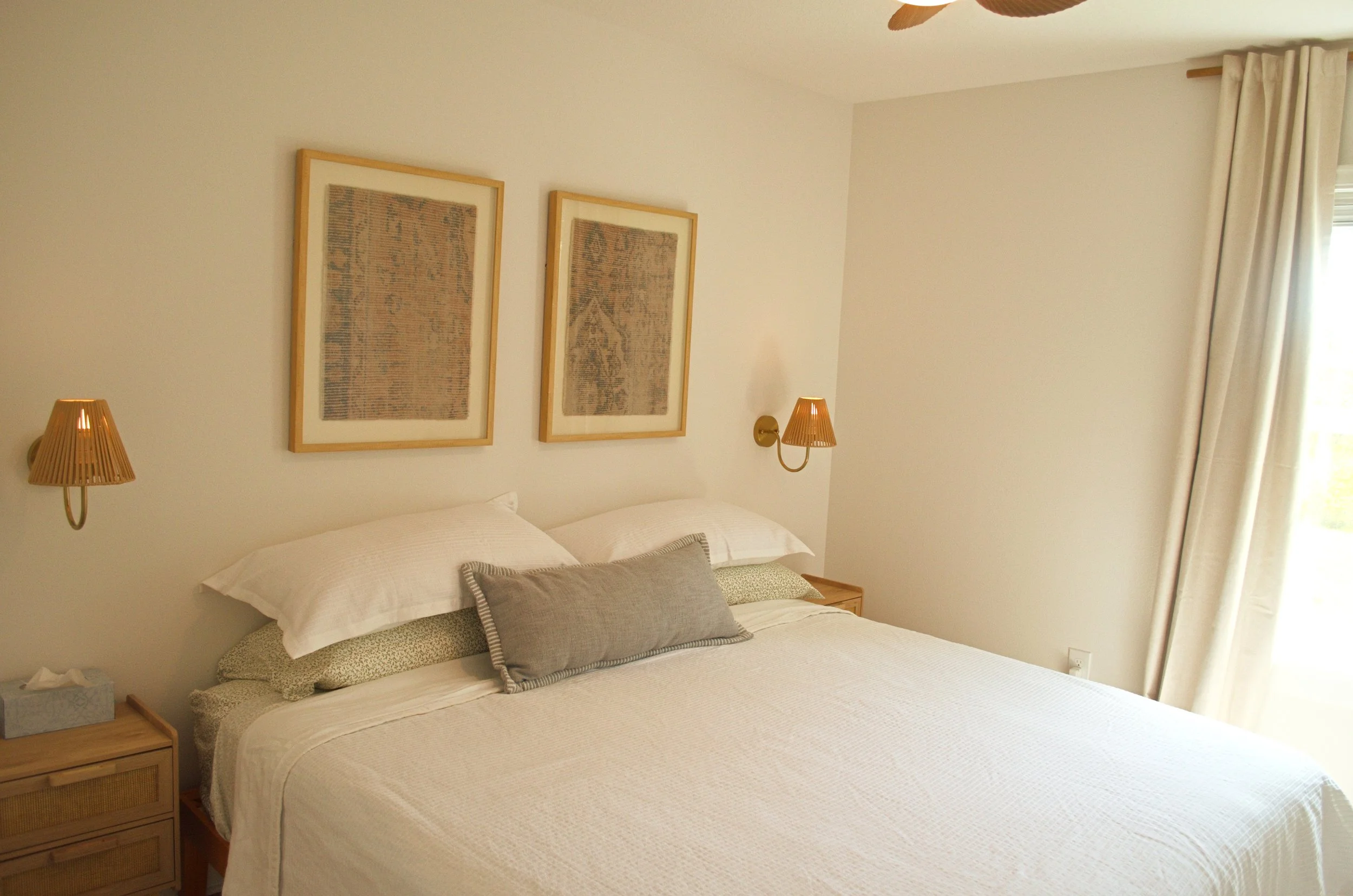 A cozy bedroom with a bed, pillows, lamps on the wall, framed artworks above the bed, and curtains on the window.