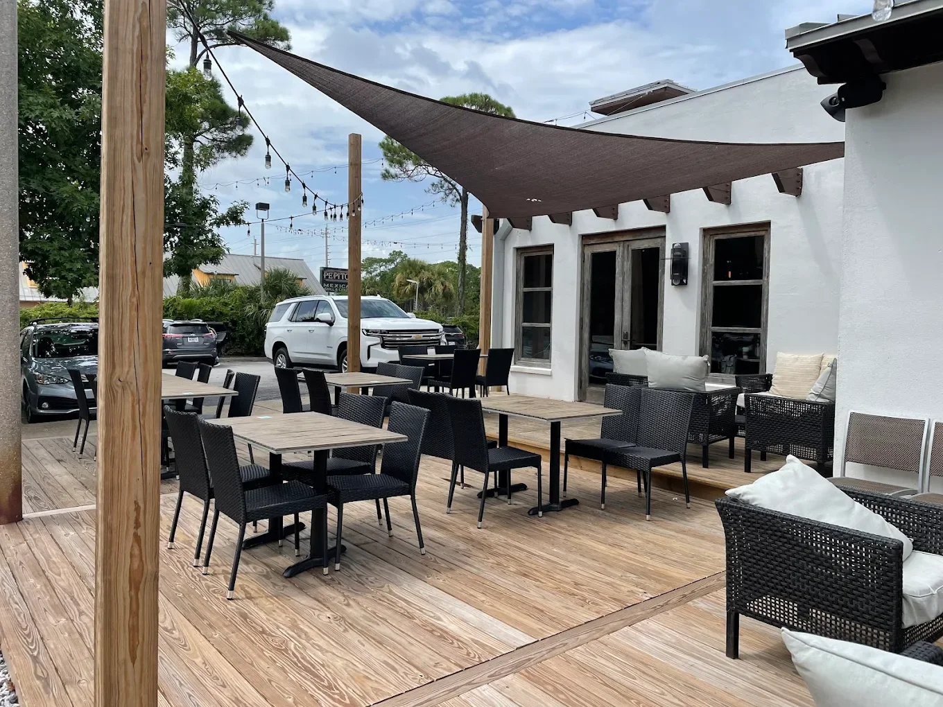 Outdoor patio with wooden flooring, black chairs, tables, cushioned black wicker seating, and string lights overhead. A white building with windows and a door, and parked cars in the background.