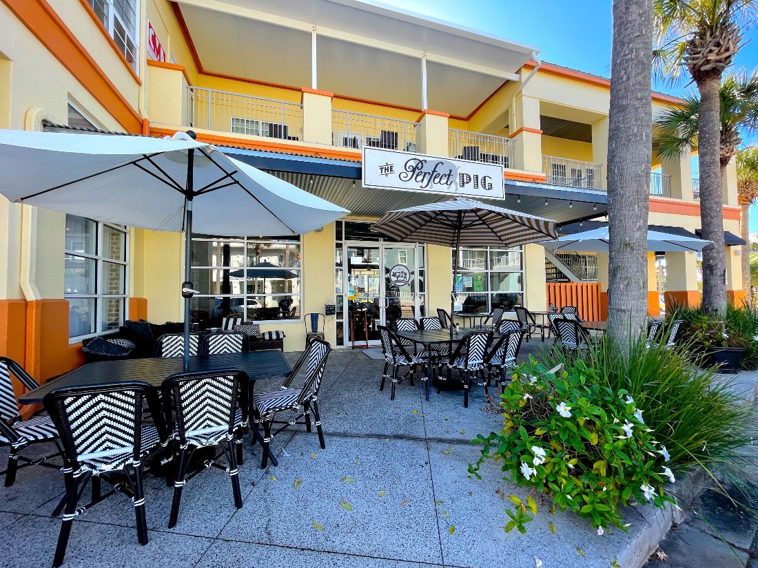 Outdoor patio seating area in front of a cafe called 'The Perfect Pig,' with tables, chairs, umbrellas, and surrounding plants.