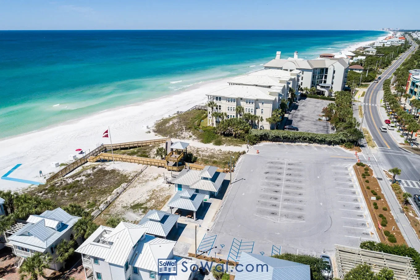 Aerial view of a beach with nearby parking lot, oceanfront buildings, and a road lined with trees.
