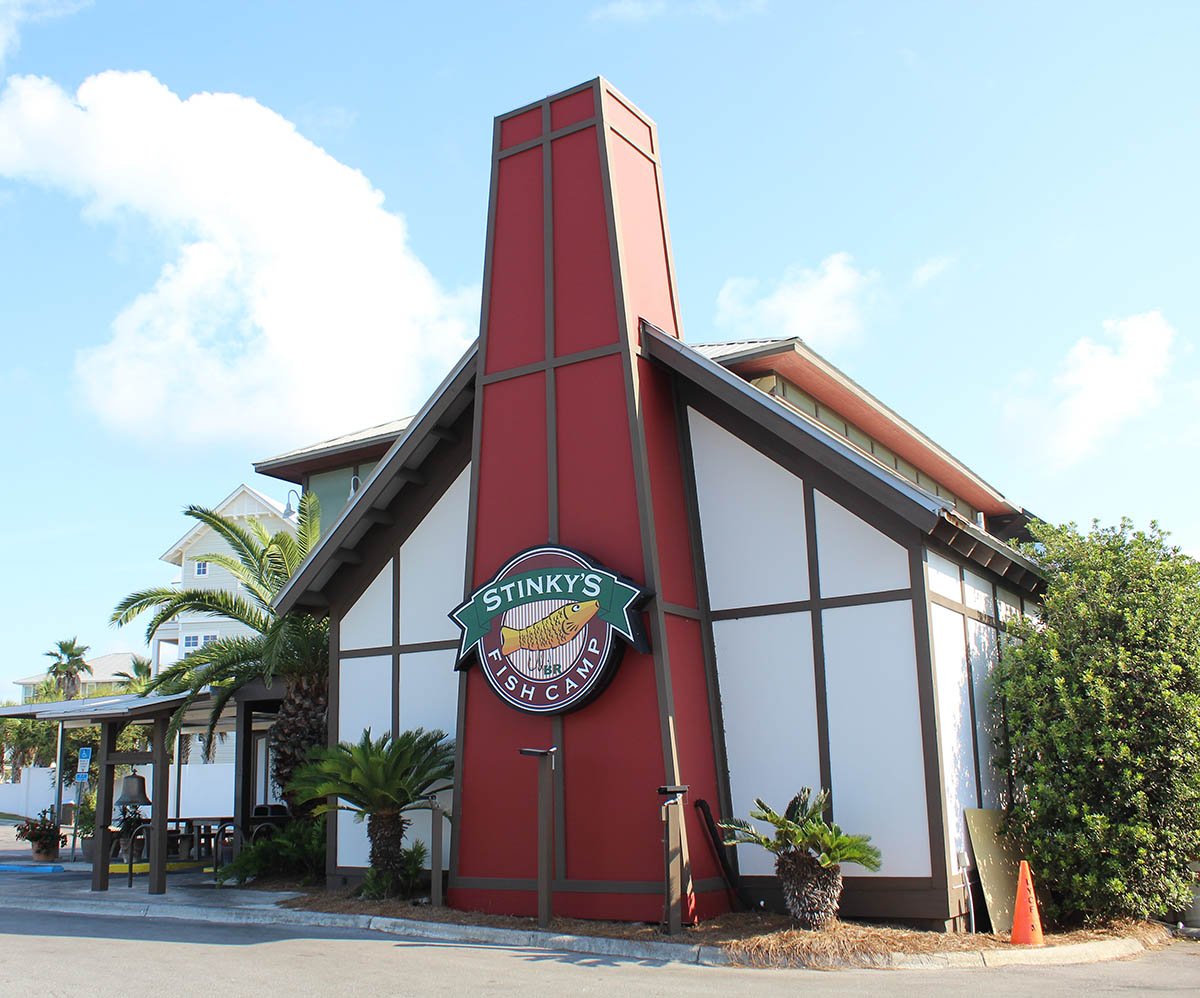 Exterior of Stinky's Fish Camp restaurant with a distinctive red and white building, tropical plants, and a blue sky with clouds.