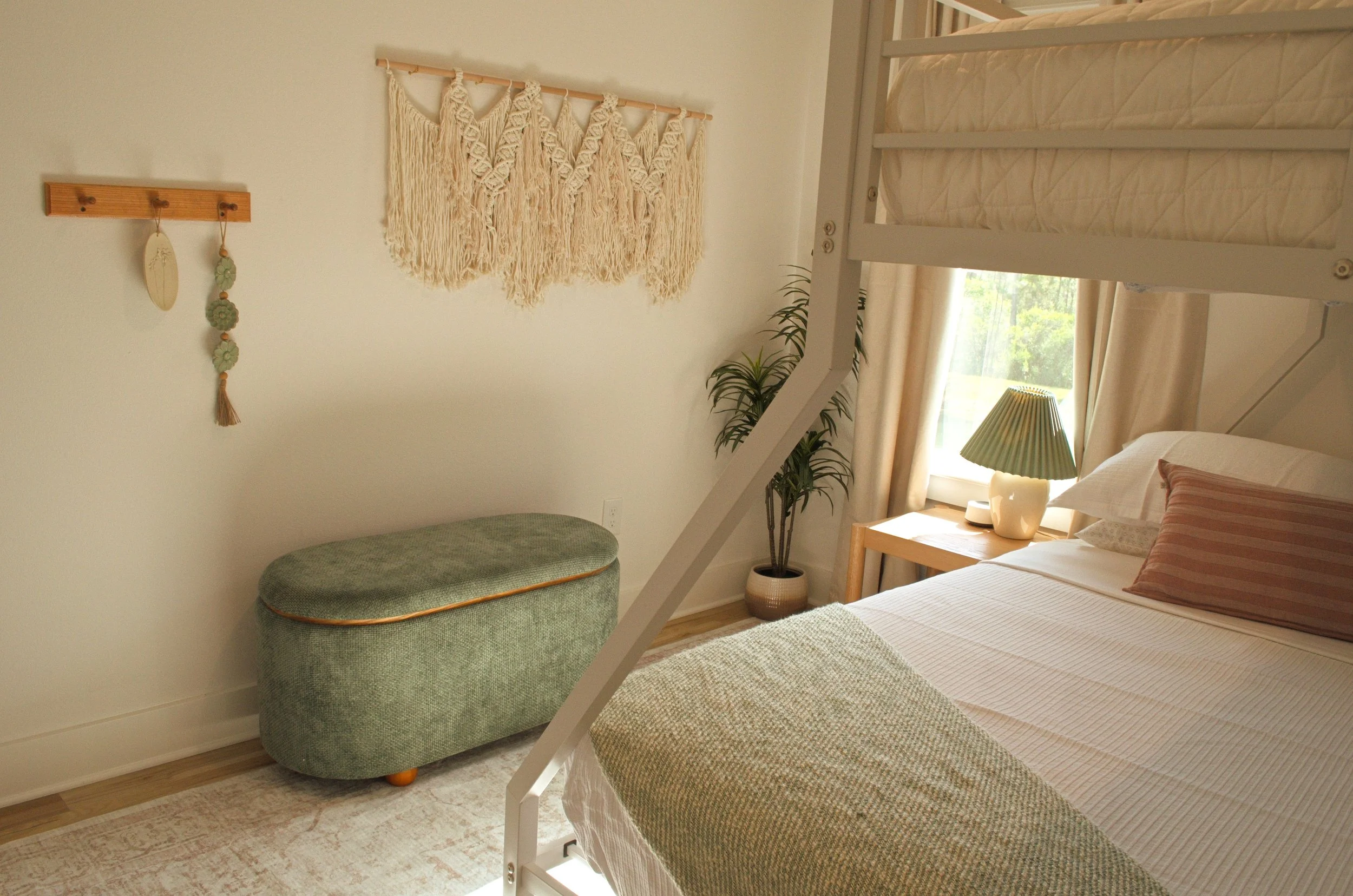 A bedroom with a bunk bed, a macrame wall hanging, a small green upholstered bench, potted plant, and a window with curtains.
