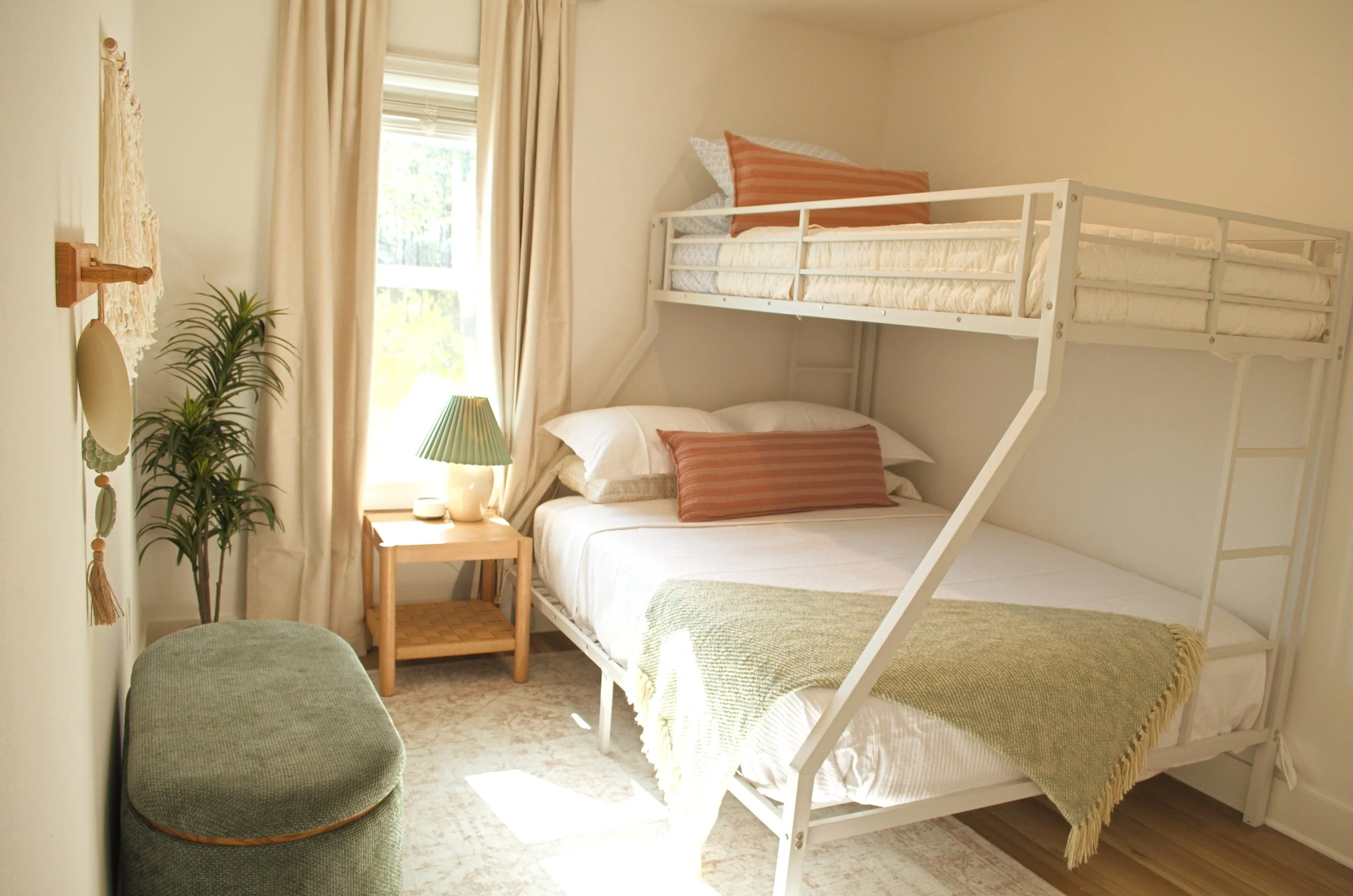 A bedroom with a bunk bed, a small wooden side table, a table lamp, a green bench, and a potted plant near a window with cream curtains, bathed in natural light.