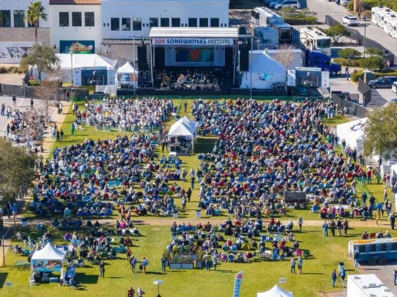 An outdoor music festival with a large crowd seated on grass in front of a stage labeled 'Songwriters Festival.' There are tents, vendors, and parked cars surrounding the event area.