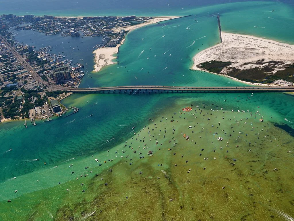 Aerial view of a coastal area showing a bridge over water, a city on the left, and a beach on the right, with boats in the water and greenish areas indicating algae or seaweed.