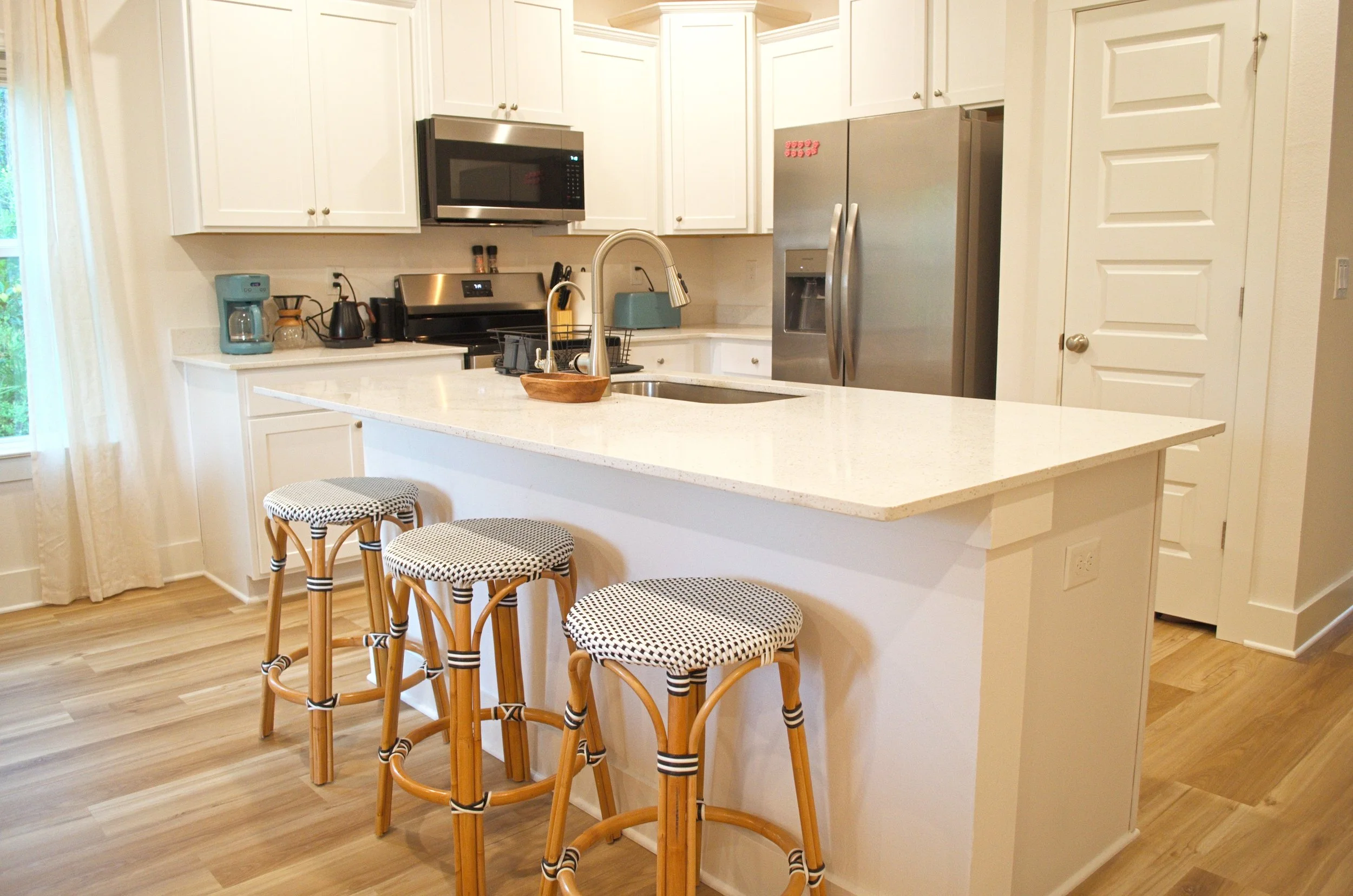 Modern kitchen with white cabinets, stainless steel appliances including a microwave, oven, and refrigerator. A kitchen island with a sink and three woven rattan barstools with black and white patterned seats. Light wood flooring and a window with wh
