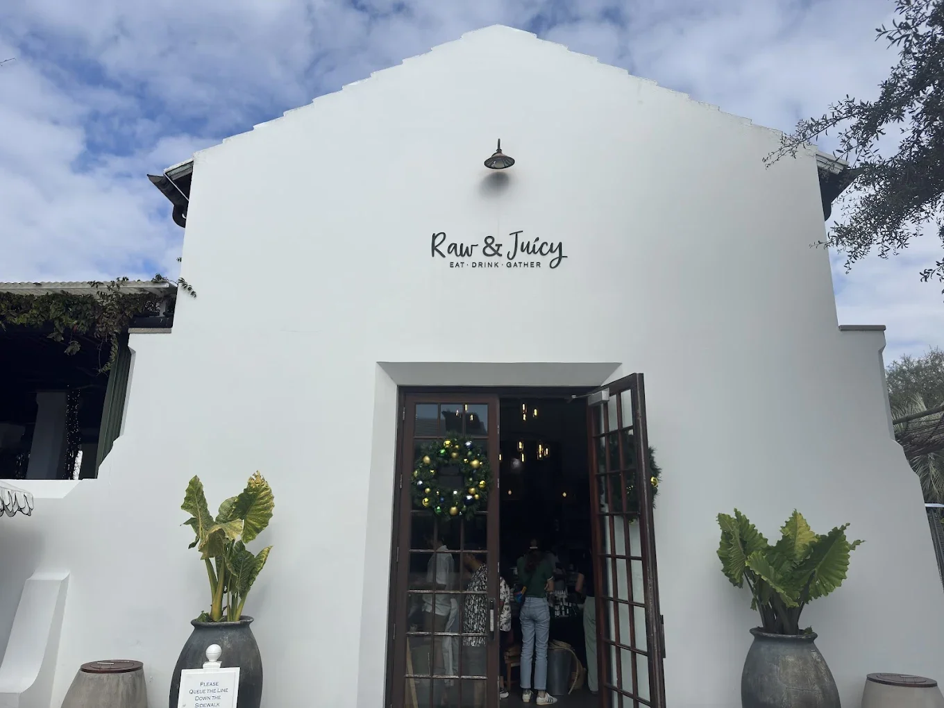 Exterior of a white building with the sign 'Raw & Juicy' and the tagline 'Eat, Drink, Gather' above the entrance. There are potted plants on either side of the doorway, and a Christmas wreath decorates the glass door, indicating a holiday season.