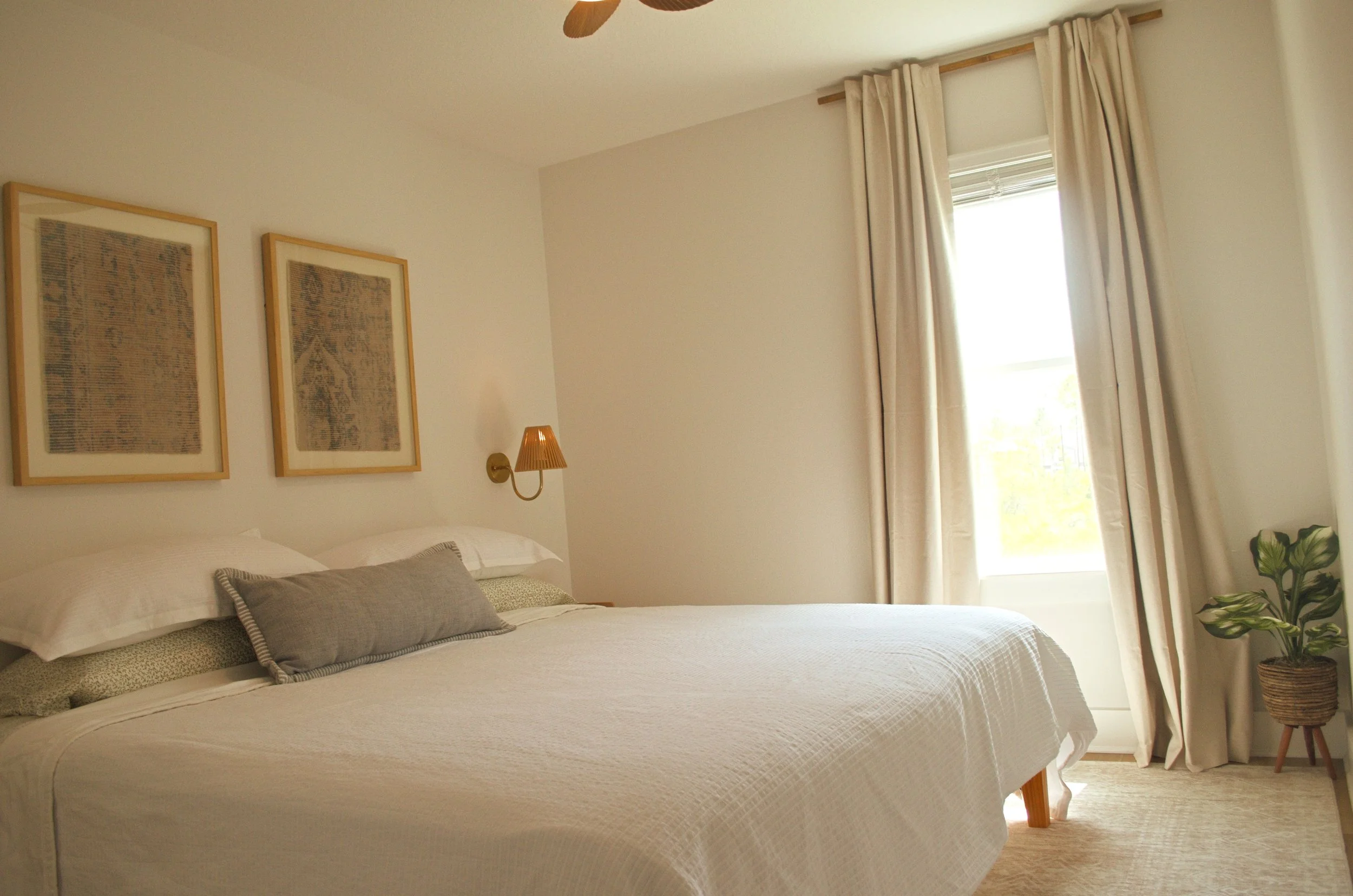 A bedroom with a bed, pillows, and beige bedding. There are three framed artworks above the bed, a bedside lamp, a window with beige curtains, and a potted plant in a woven basket. The room has a light color palette and natural light.