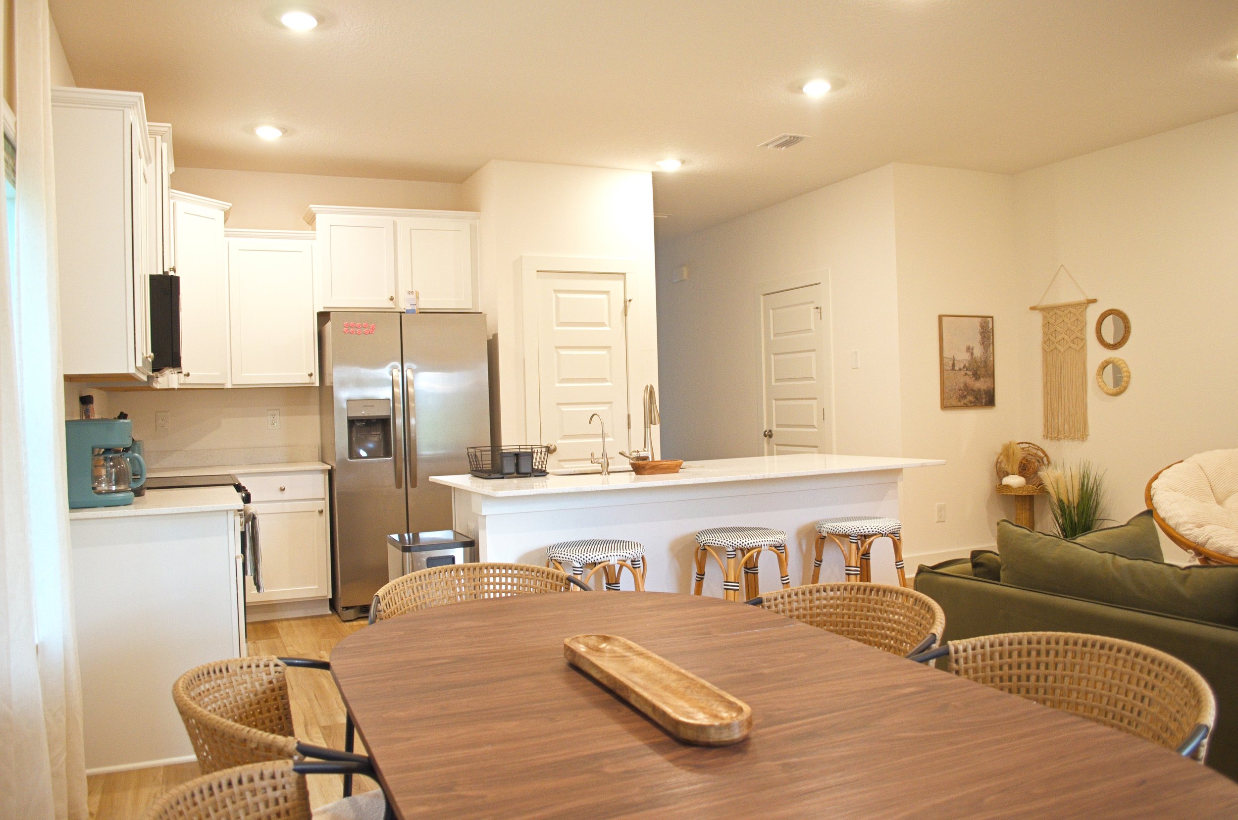 A kitchen and dining area with white cabinets, stainless steel refrigerator, a sink, and a wooden table with wicker chairs. There's a green sofa, wall art, and decorative hanging on the wall.