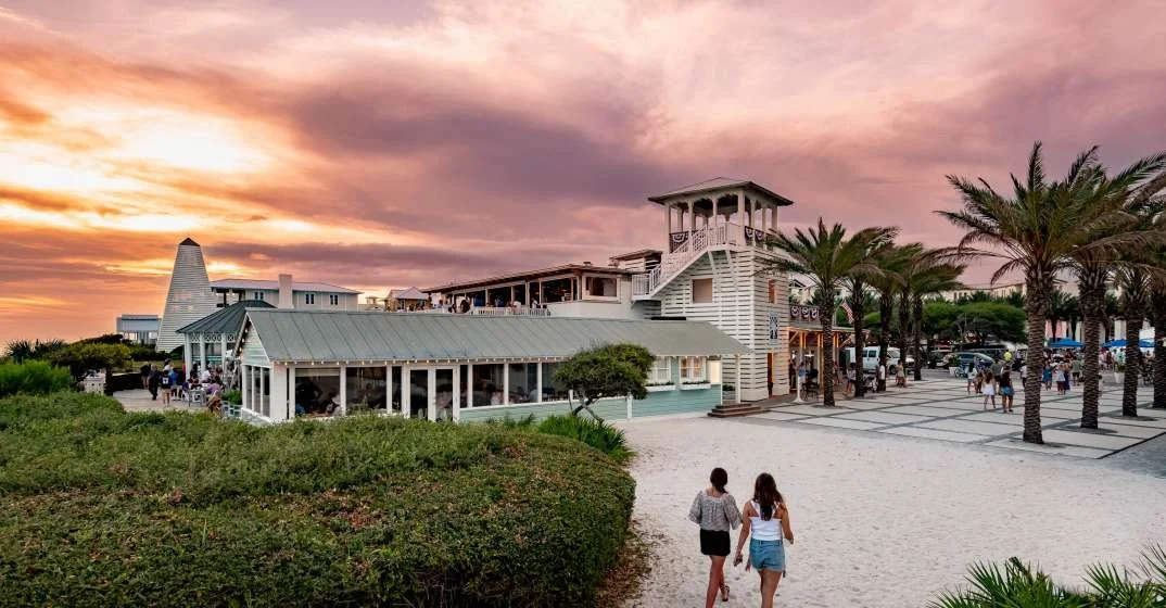 People walking toward a beachside restaurant with palm trees and a sunset sky.