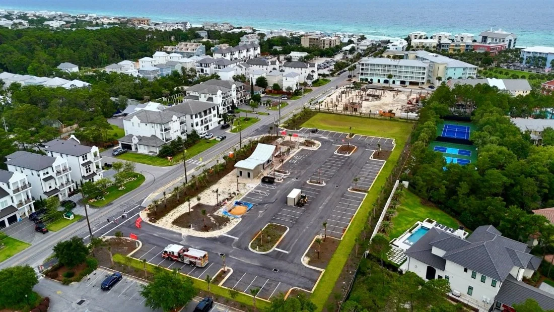 Aerial view of a coastal residential neighborhood with newly constructed apartment buildings, a rec center with a swimming pool and a playground, and tennis courts surrounded by trees and greenery.
