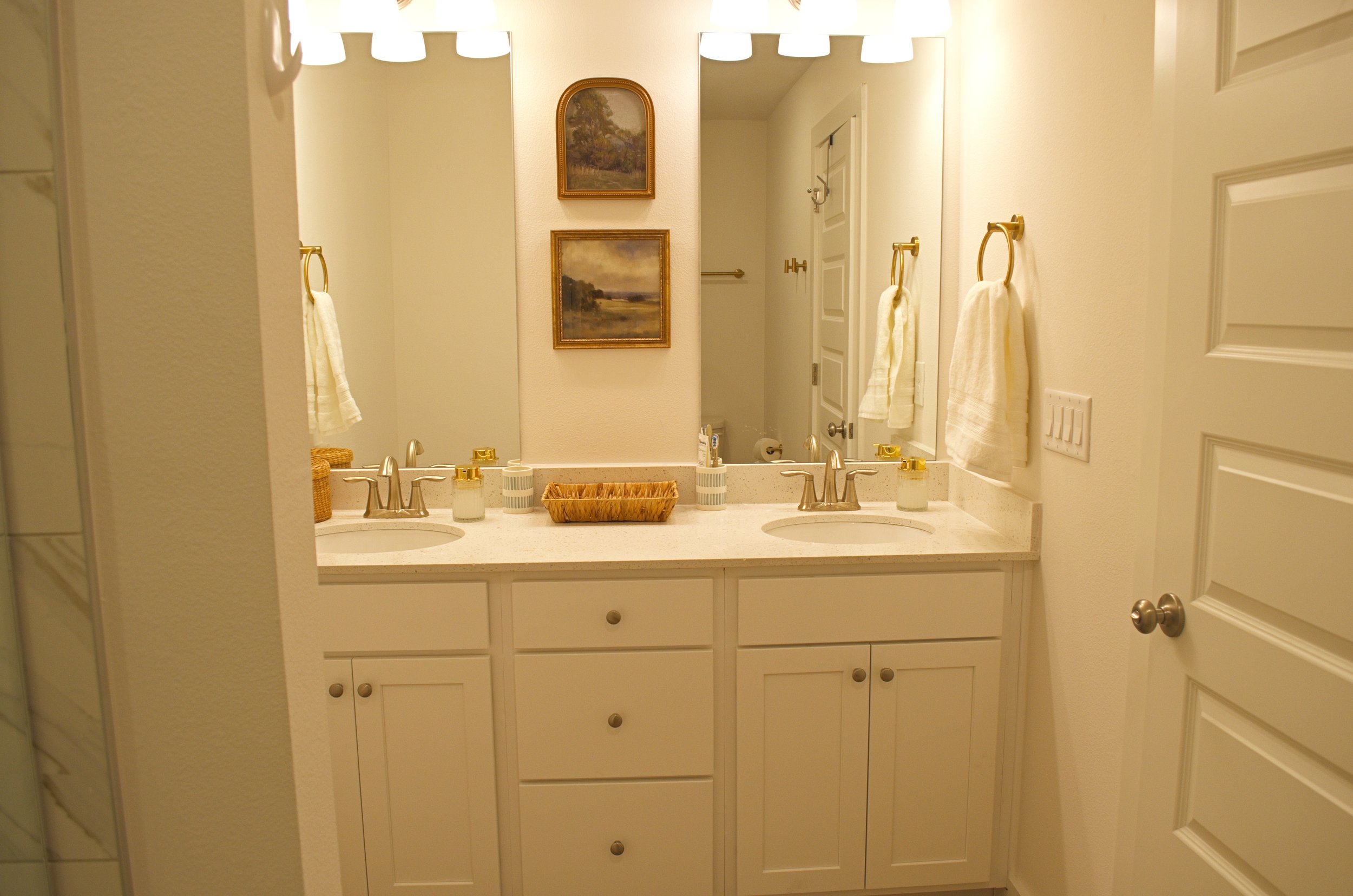 Double bathroom vanity with two sinks, decorated with soap dispensers, small buckets, and a wicker tray, reflected in a mirror. The wall has two art paintings. There are towel rings with towels on each side of the mirror, and a door to the right.