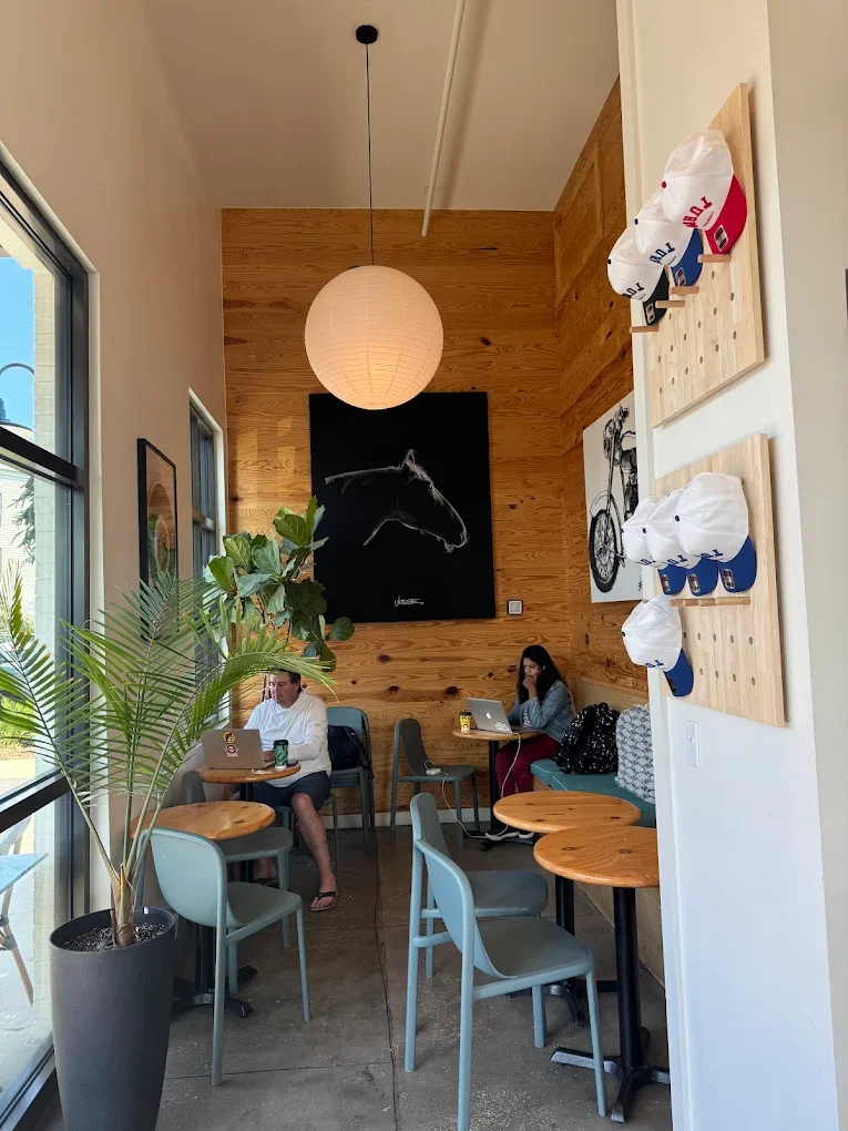 Interior of a cafe with wooden accent wall, two people working on laptops, large window with plant nearby, and baseball caps hanging on a wall-mounted pegboard.