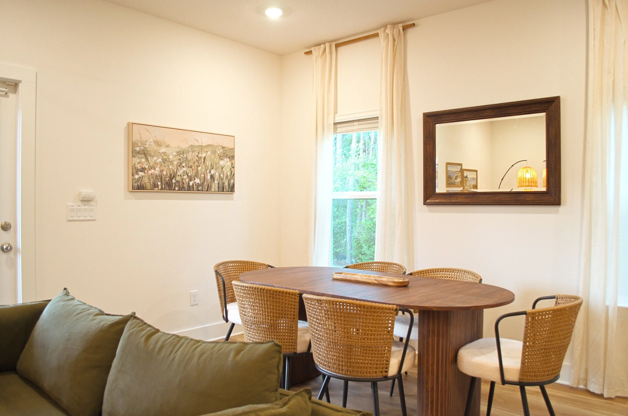A living room with a dining area, featuring a wooden round table with several wicker chairs, a large mirror on the wall, a painting of flowers, large windows with white curtains, and a view of trees outside.