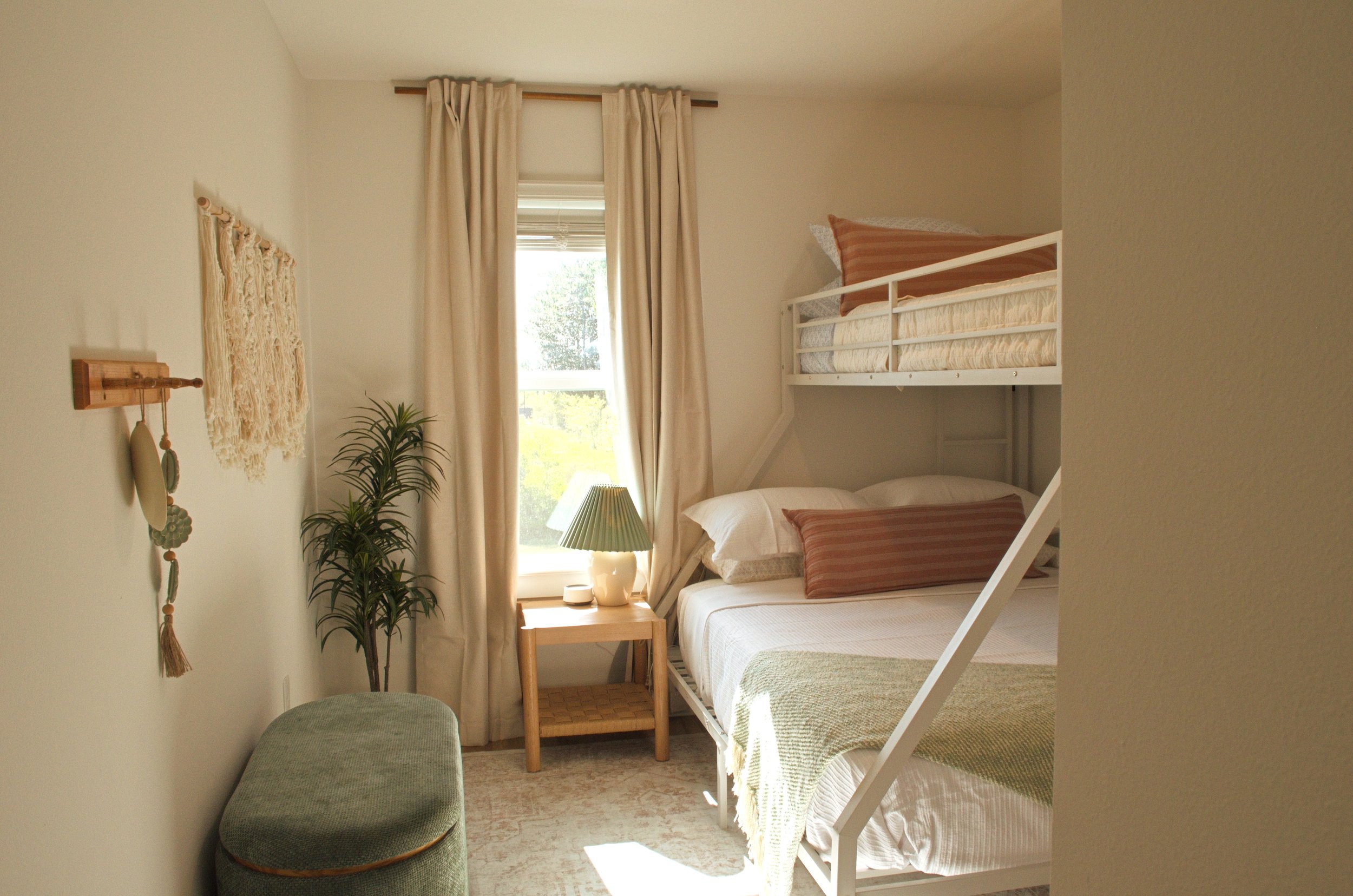 A cozy bedroom with two stacked beds in white frames, beige and brown pillows, a nightstand with a lamp, a window with cream curtains, a green plant, a macrame wall hanging, and a green upholstered bench at the foot of the bed.