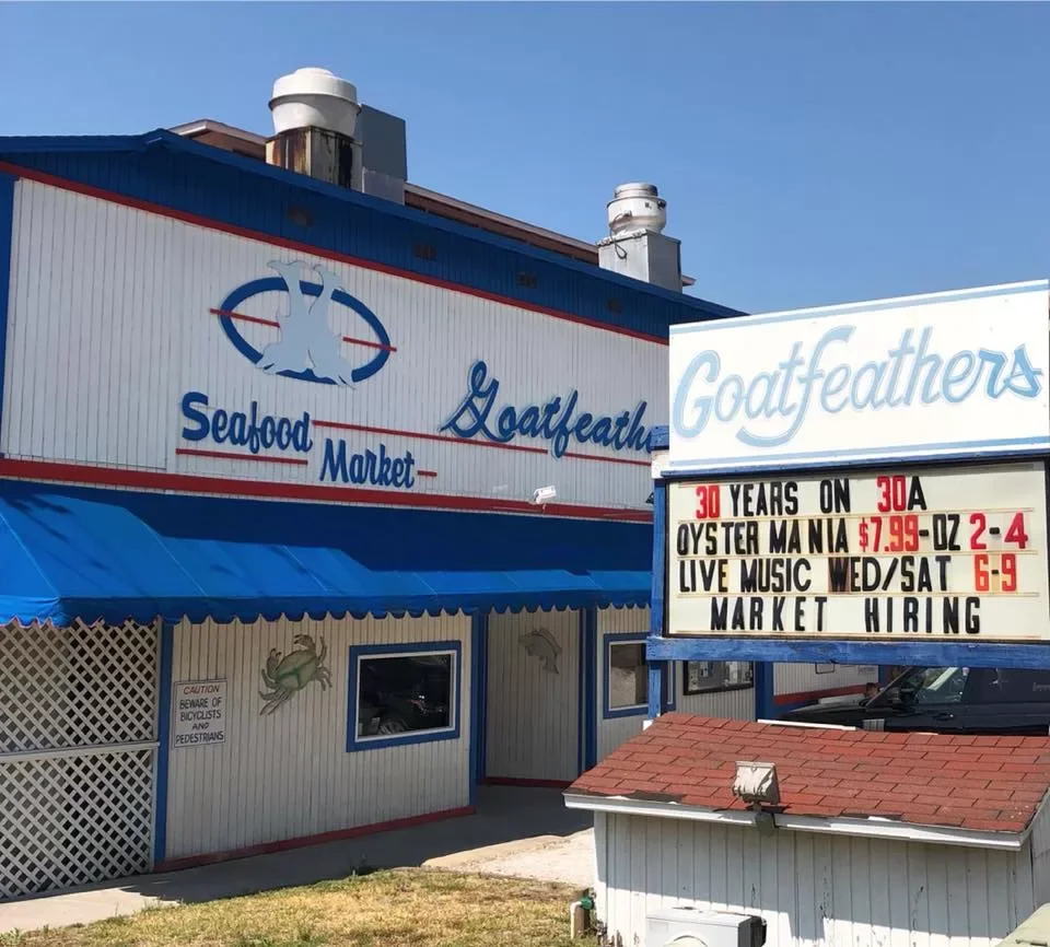 Exterior of a seafood market with a sign that reads 'Goatfeathers.' The sign mentions 30 years on 30A, oyster mania for $7.99 per ounce, live music on Wednesday and Saturday from 6 to 9, and a market selling seafood.