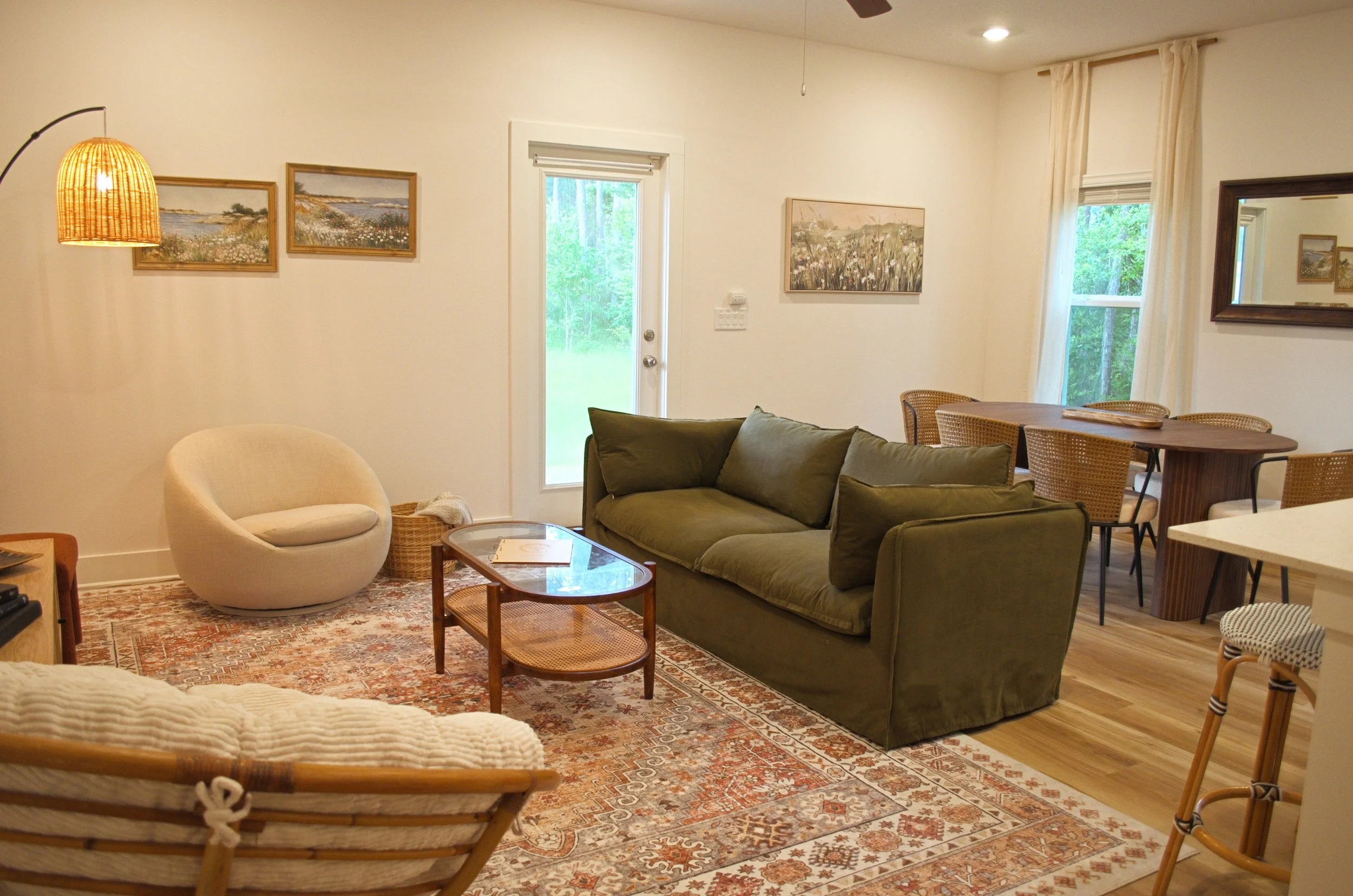 Living room with a green sofa, a cream armchair, a wooden coffee table, a patterned rug, and framed landscape paintings on white walls. There are large windows with white curtains and a door leading outside.