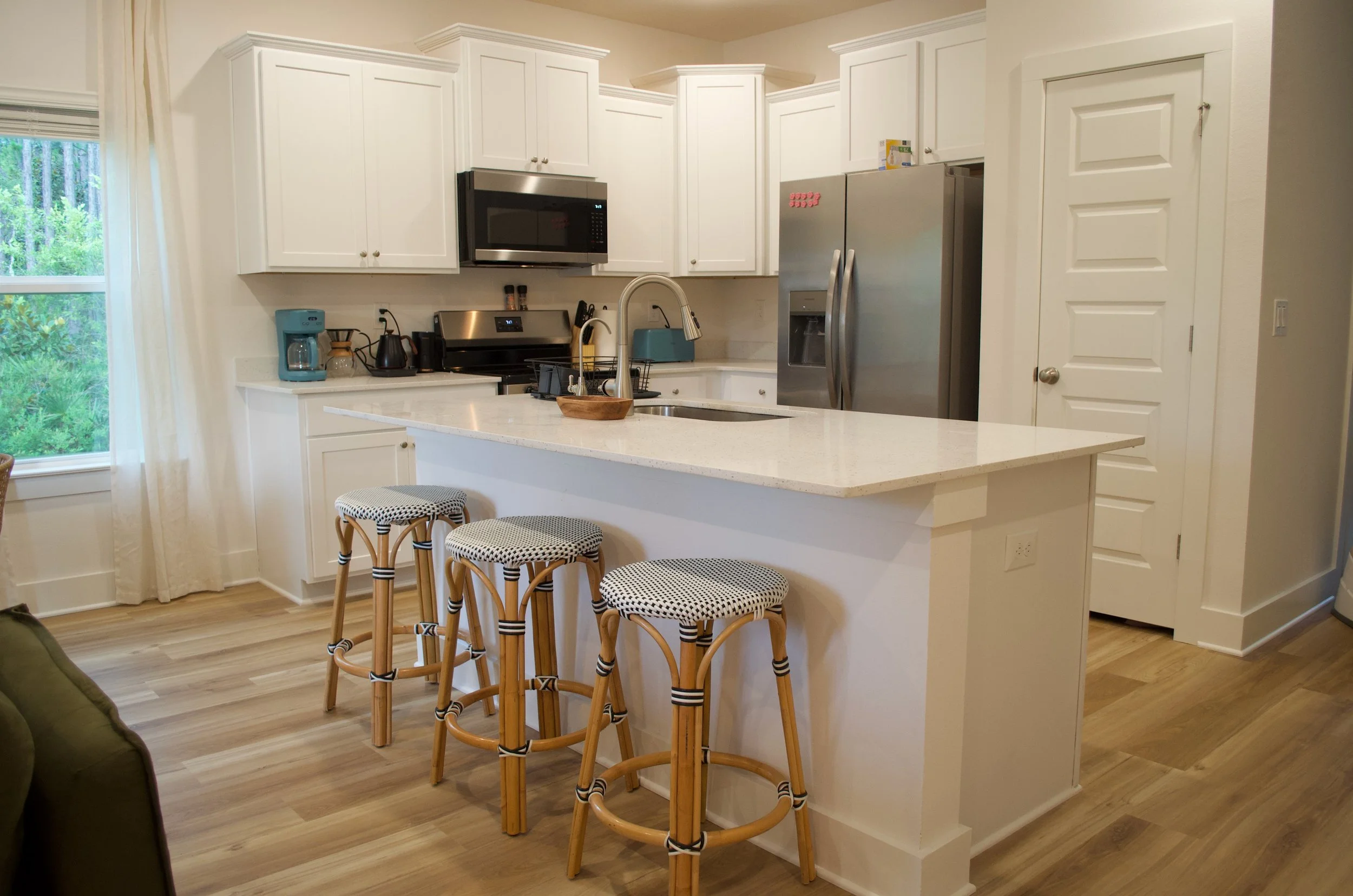 A modern kitchen with white cabinets, stainless steel appliances, a kitchen island with a white countertop, four rattan stools with black-and-white patterned cushions, and a window with white curtains overlooking trees.