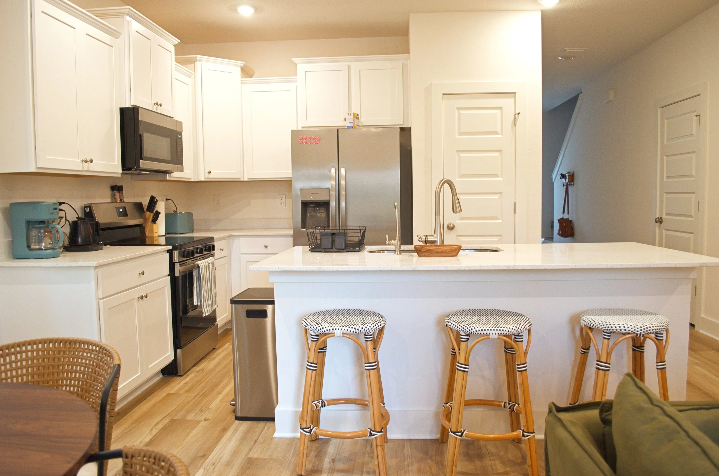 A modern, bright kitchen with white cabinets, stainless steel appliances, a kitchen island with bar stools, and a doorway leading to a hallway.