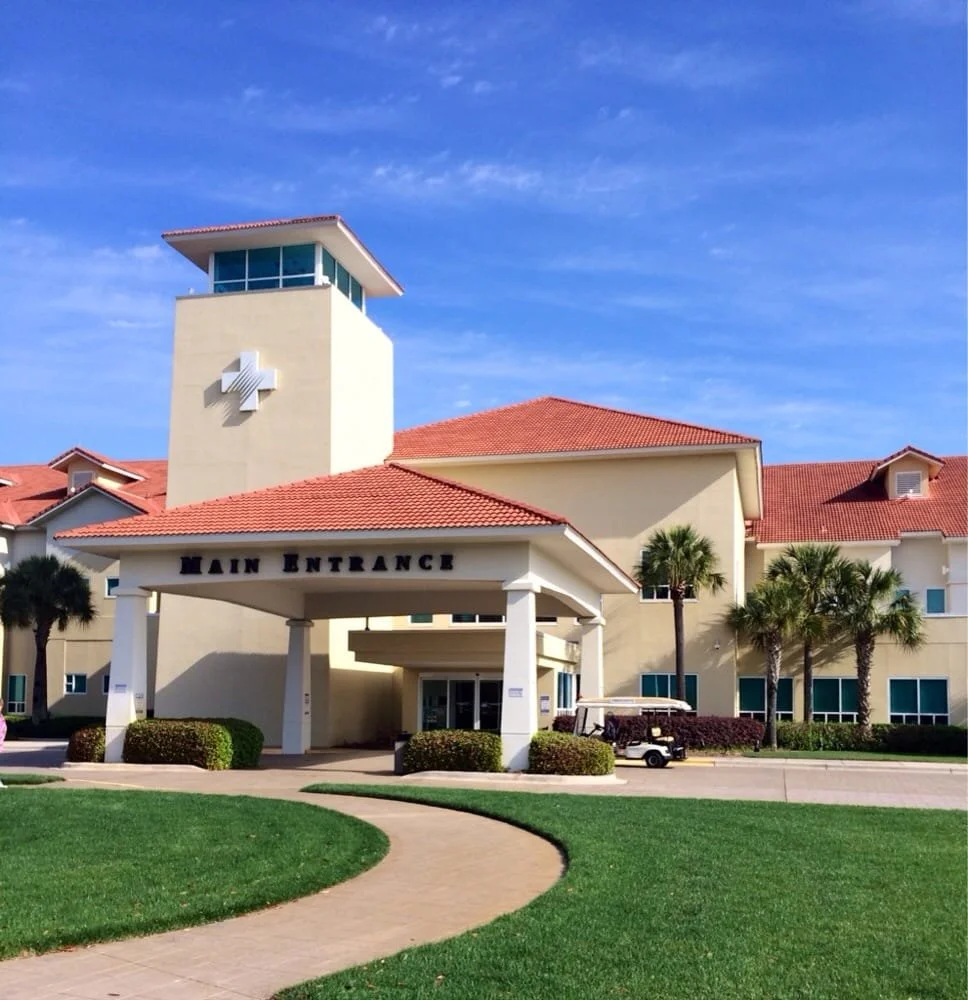 A hospital or medical facility with a prominent cross symbol on a tower and a main entrance labeled 'Main Entrance'. The building has red-tiled roofs, palm trees, and a golf cart parked outside. The sky is clear and blue.