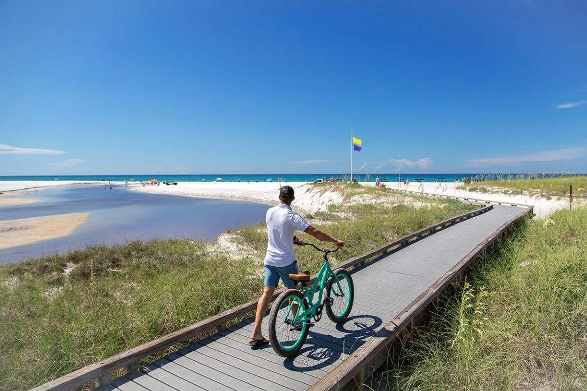 A man with a bicycle on a wooden boardwalk near the beach, with a blue sky, white sand, and ocean in the background.