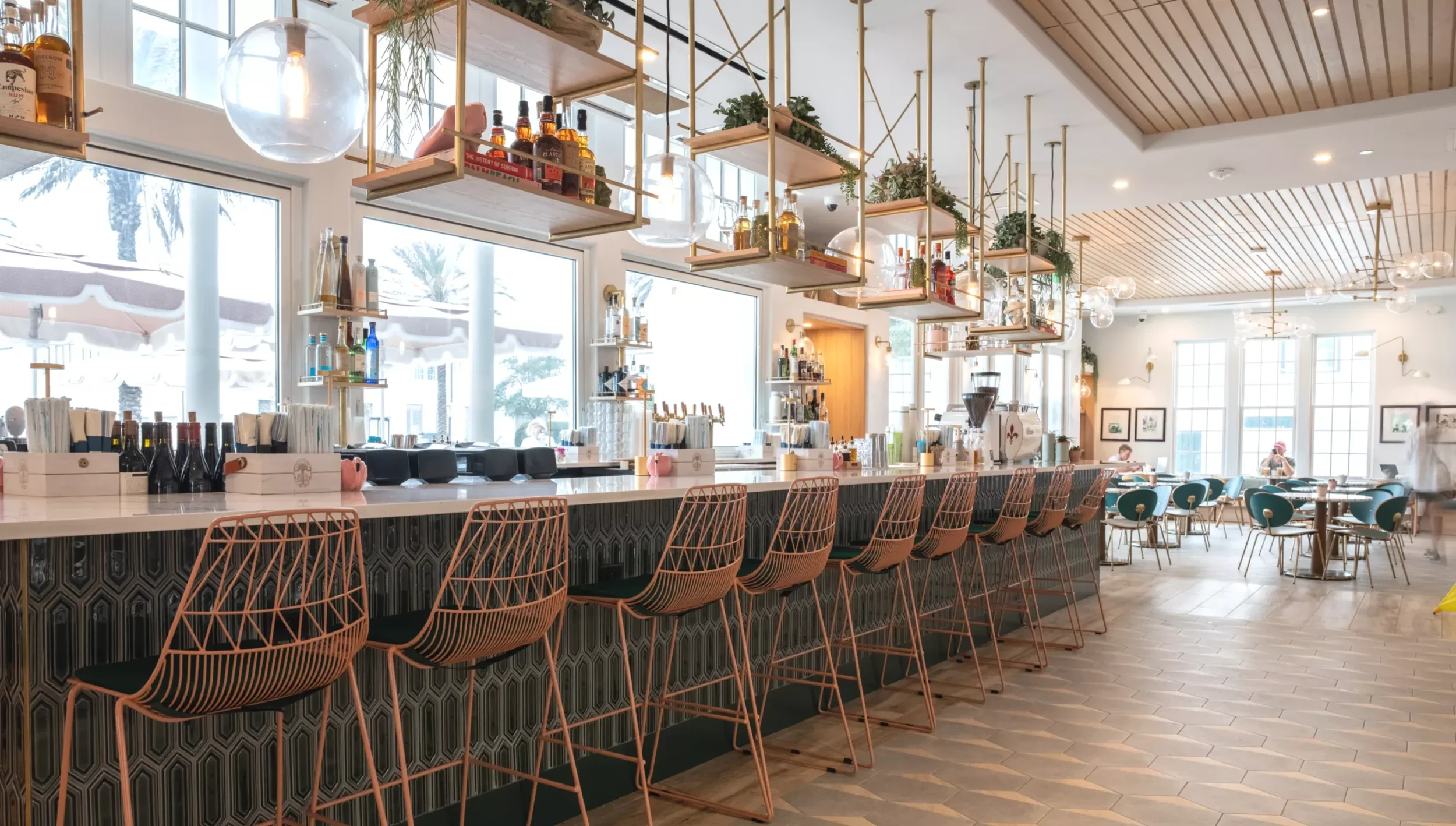 Inside a bright, modern restaurant or cafe with a long bar counter and seating. The bar has pink wire chairs in front of a white countertop with various bottles and drink supplies. Overhead, decorative shelving holds bottles, books, and greenery, with large windows allowing natural light. The ceiling features wooden slats and hanging globe lights.