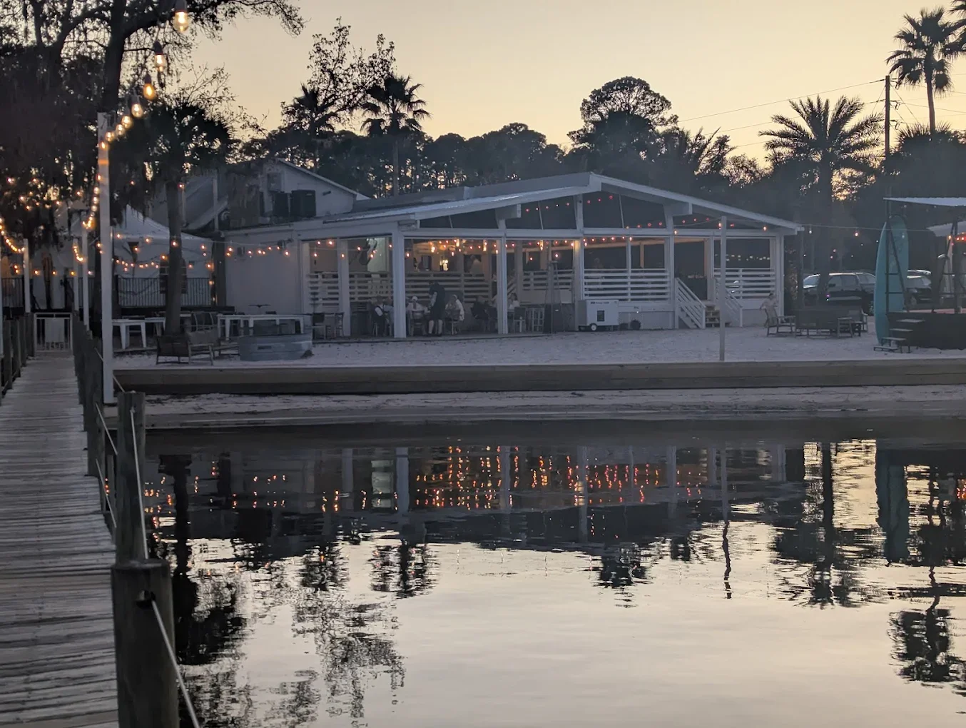 Sunset at a lakeside restaurant with string lights on a wooden deck and a structure for dining, reflecting on the calm water.