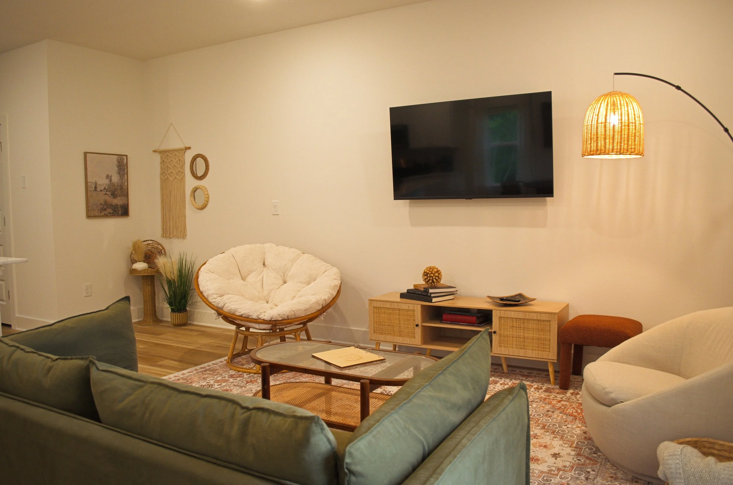 Living room with a green sofa, a rattan chair with white cushion, a wooden coffee table with books, a entertainment unit with books, a wall-mounted flat screen TV, decorative mirrors, artwork, a rattan pendant lamp, and a carpet.