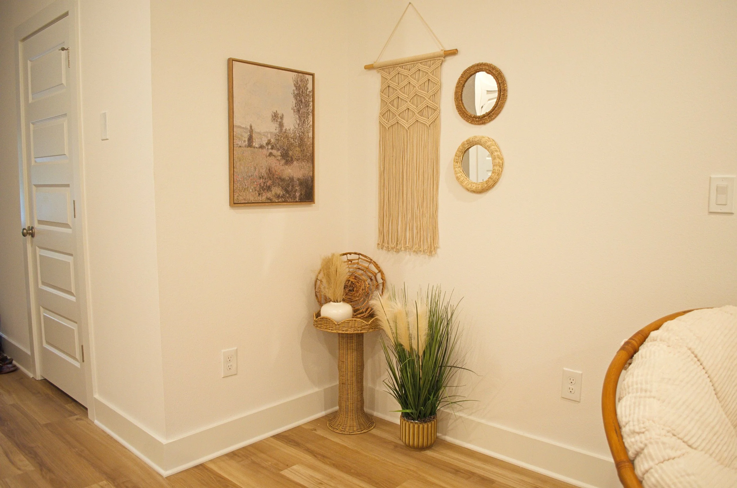 Decorative corner of a room with framed art, woven wall hangings, a wicker vase with dried plants, a potted green plant, and part of a bed with a white blanket.