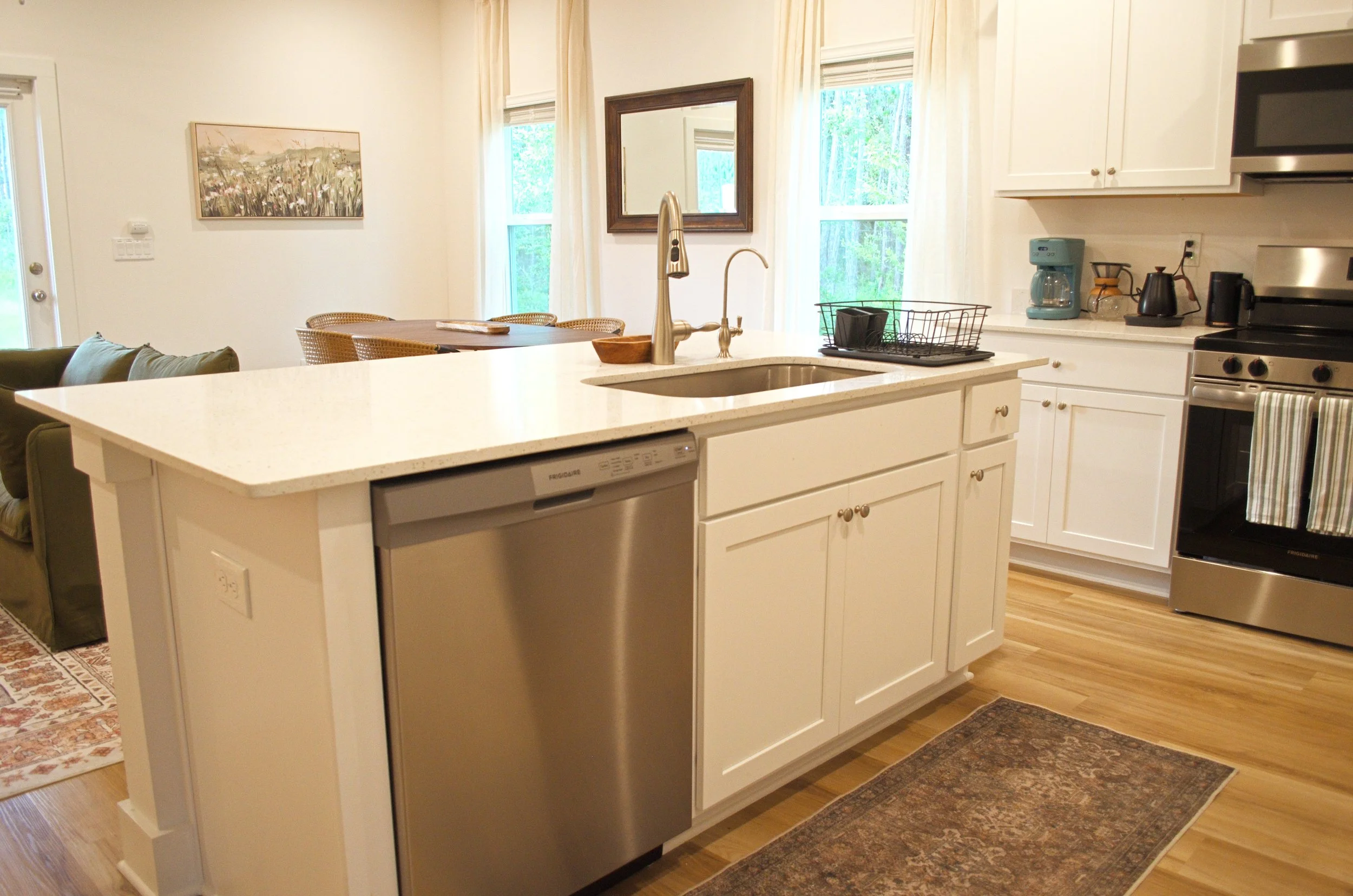 A modern kitchen with white cabinets, a white island countertop, stainless steel appliances, and a window with curtains showing greenery outside.