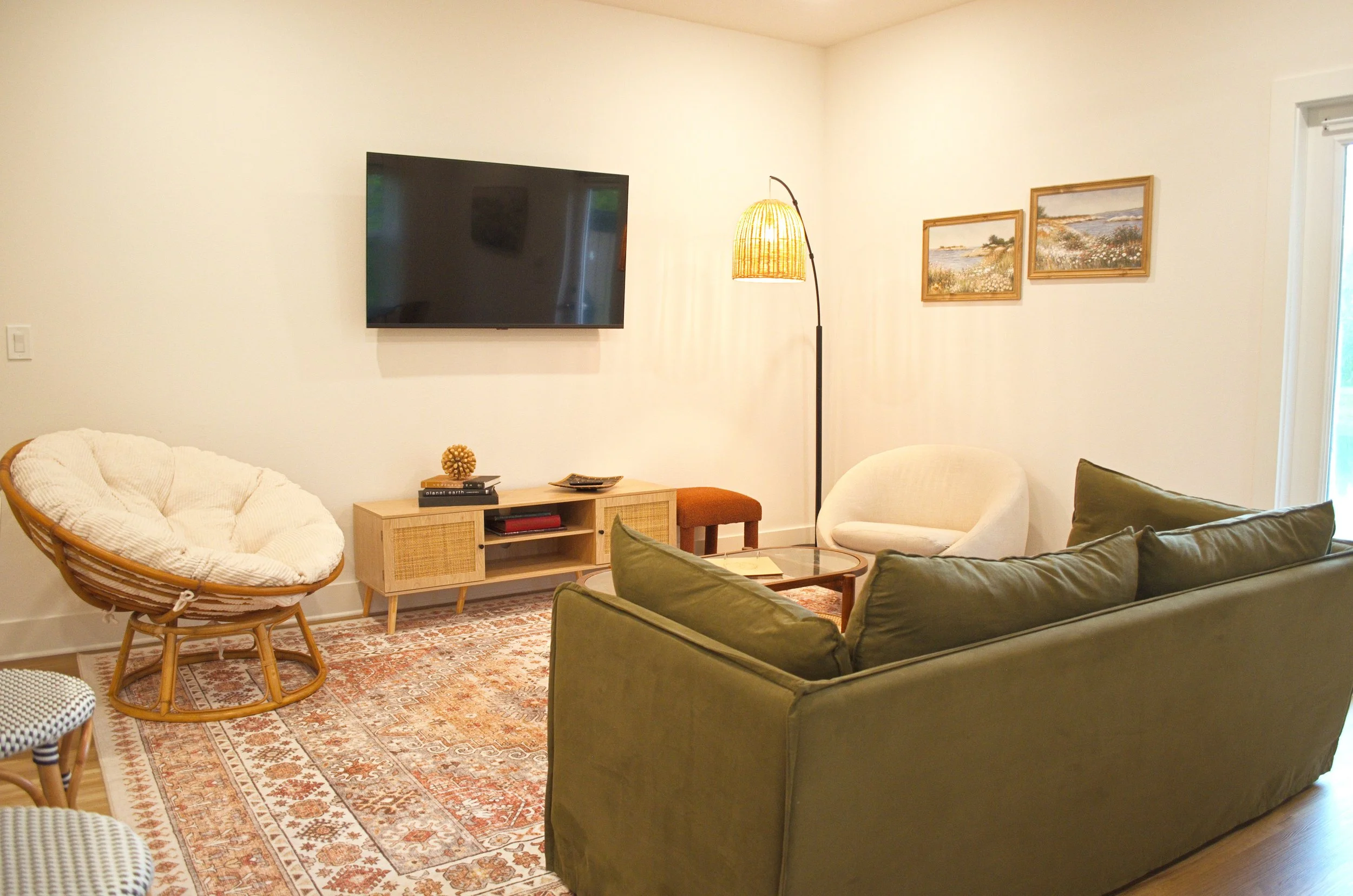 Living room with a wall-mounted TV, a wicker papasan chair, a beige armchair, a green sofa, a wooden TV stand with books and decorative items, a patterned rug, a standing lamp with a woven shade, and two landscape paintings on the wall near a window.