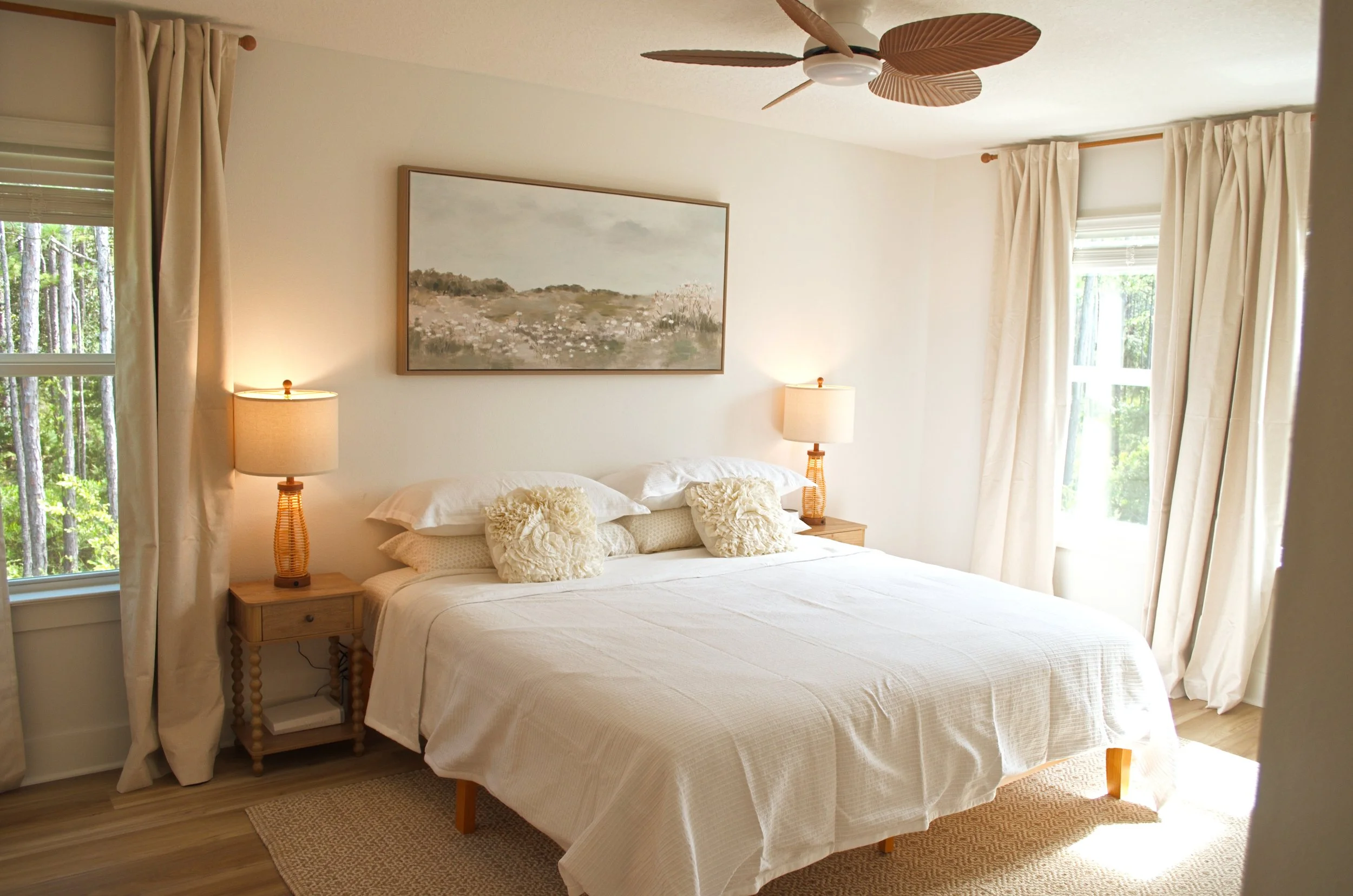 A bedroom with a neatly made bed, cream-colored bedding, and decorative pillows. There are two matching bedside tables with lamps, large windows with curtains, and a framed landscape painting on the wall. A ceiling fan with leaf-shaped blades is abov