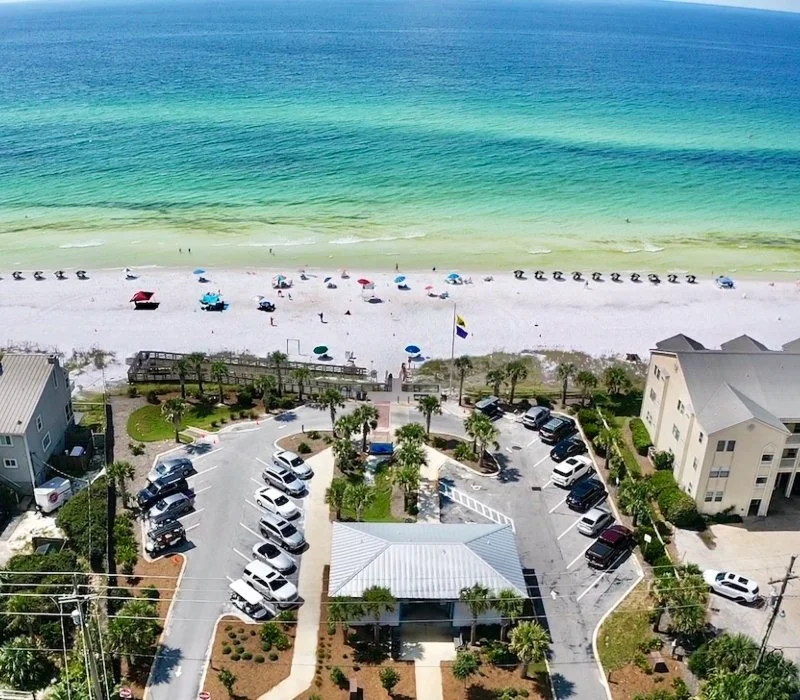 Aerial view of a beach with turquoise water, white sand, beach chairs, and umbrellas, with nearby parking lot and buildings.