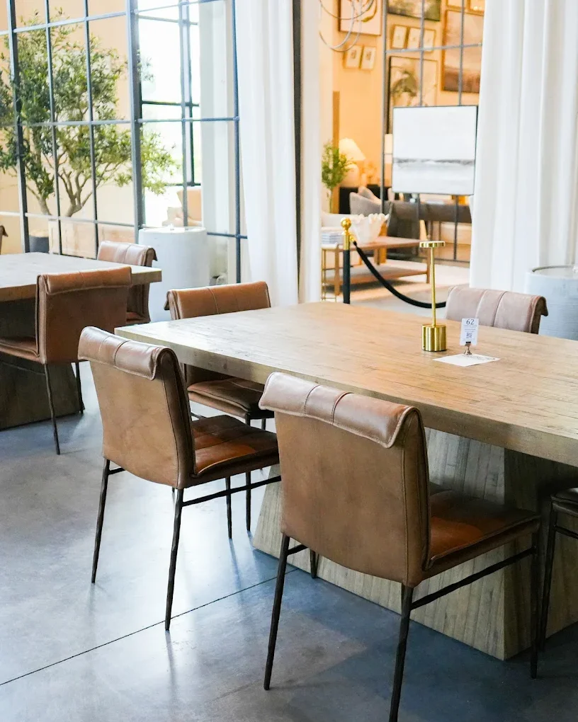 A modern dining area with a large wooden table, surrounded by six brown leather chairs. The room is bright with natural light from large windows, and there are white curtains, potted plants, framed pictures on the wall, and a small lamp on a side table visible in the background.