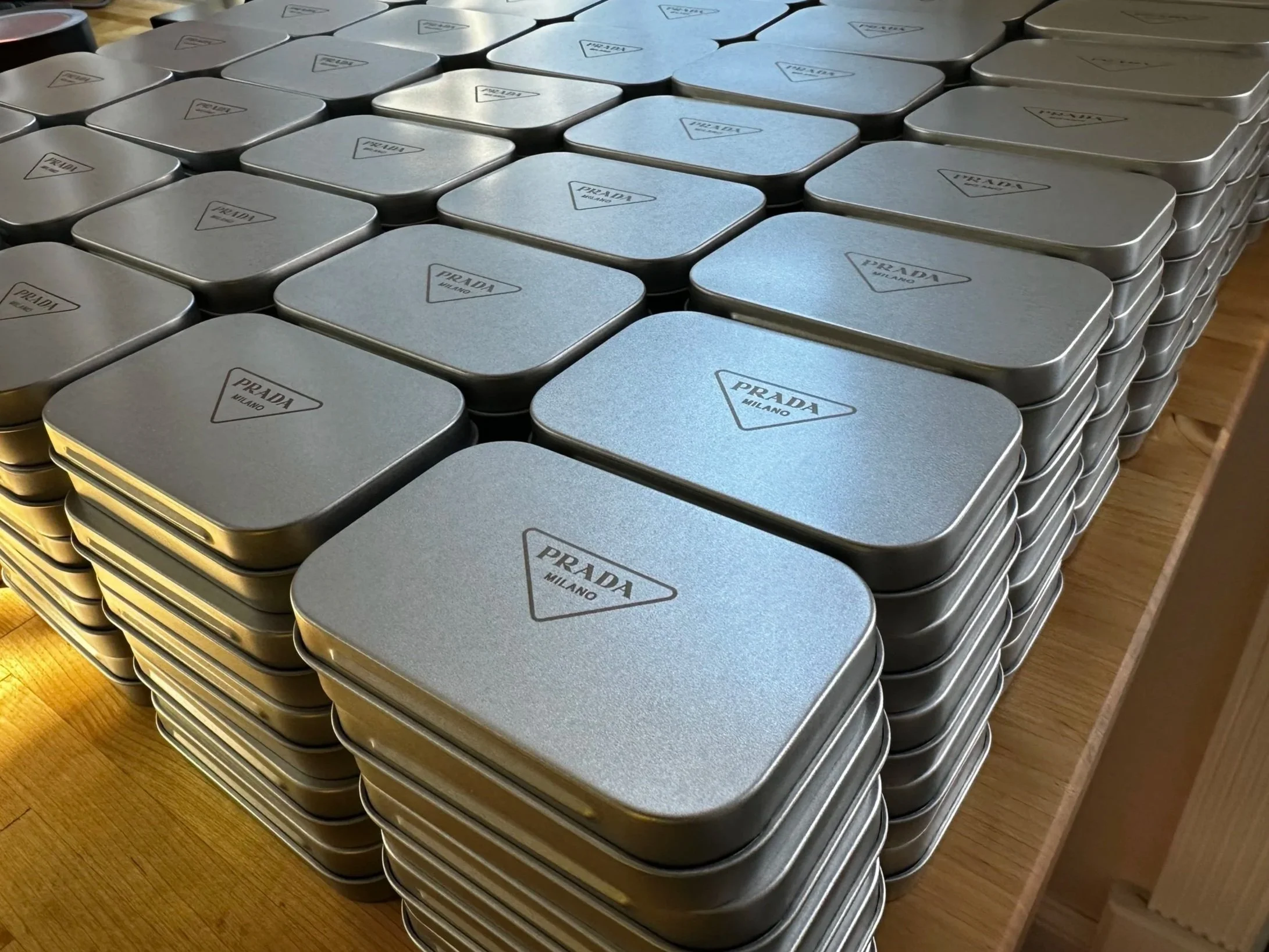 Multiple stacked silver-colored Prada Milano tins with black text and a triangle logo on top, arranged on a wooden surface.
