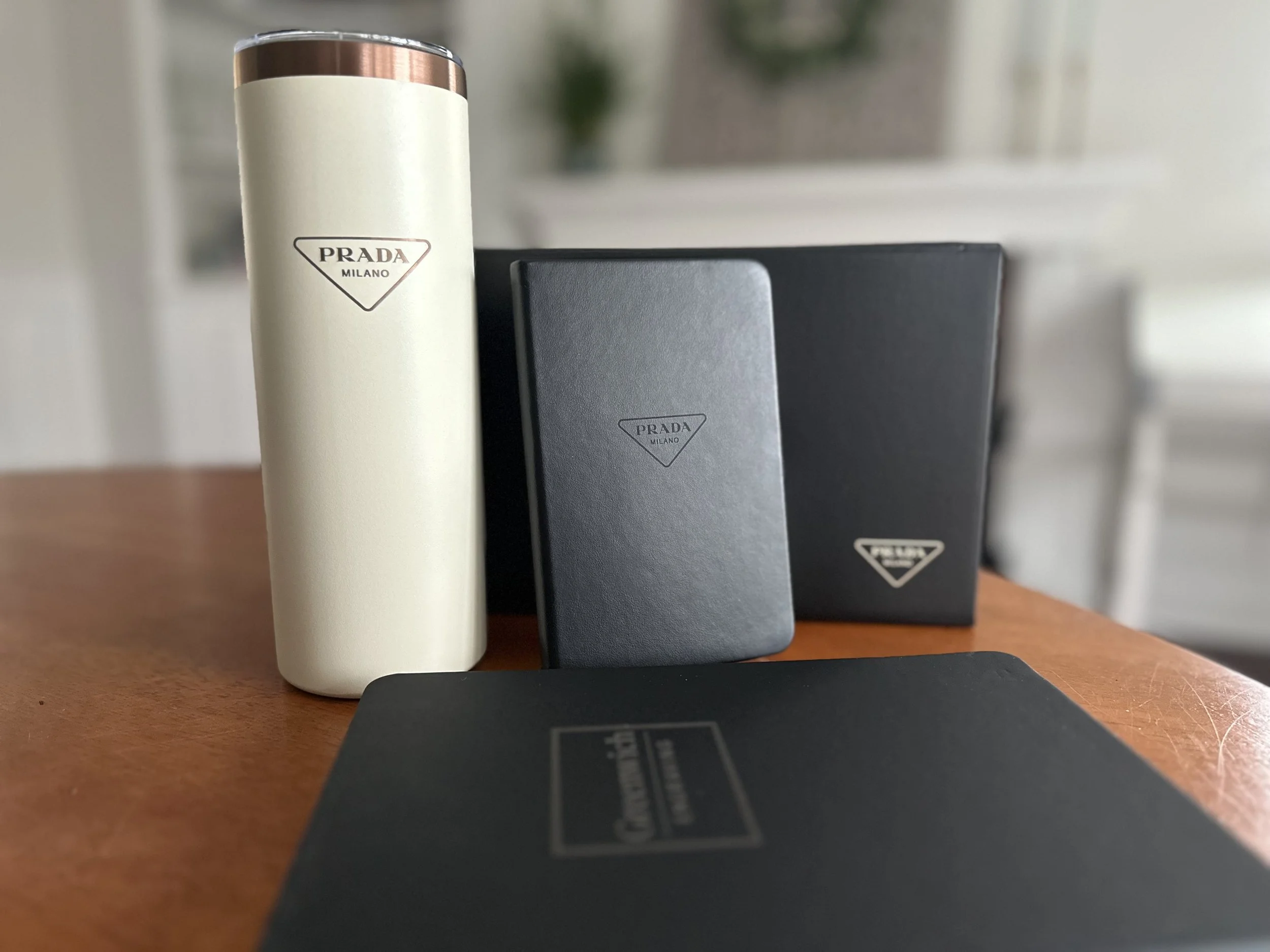 Close-up of Prada Milano branded items including a cream-colored cylindrical container with a metallic cap, a black rectangular box, and a black card or booklet on a wooden surface, with a blurred background.