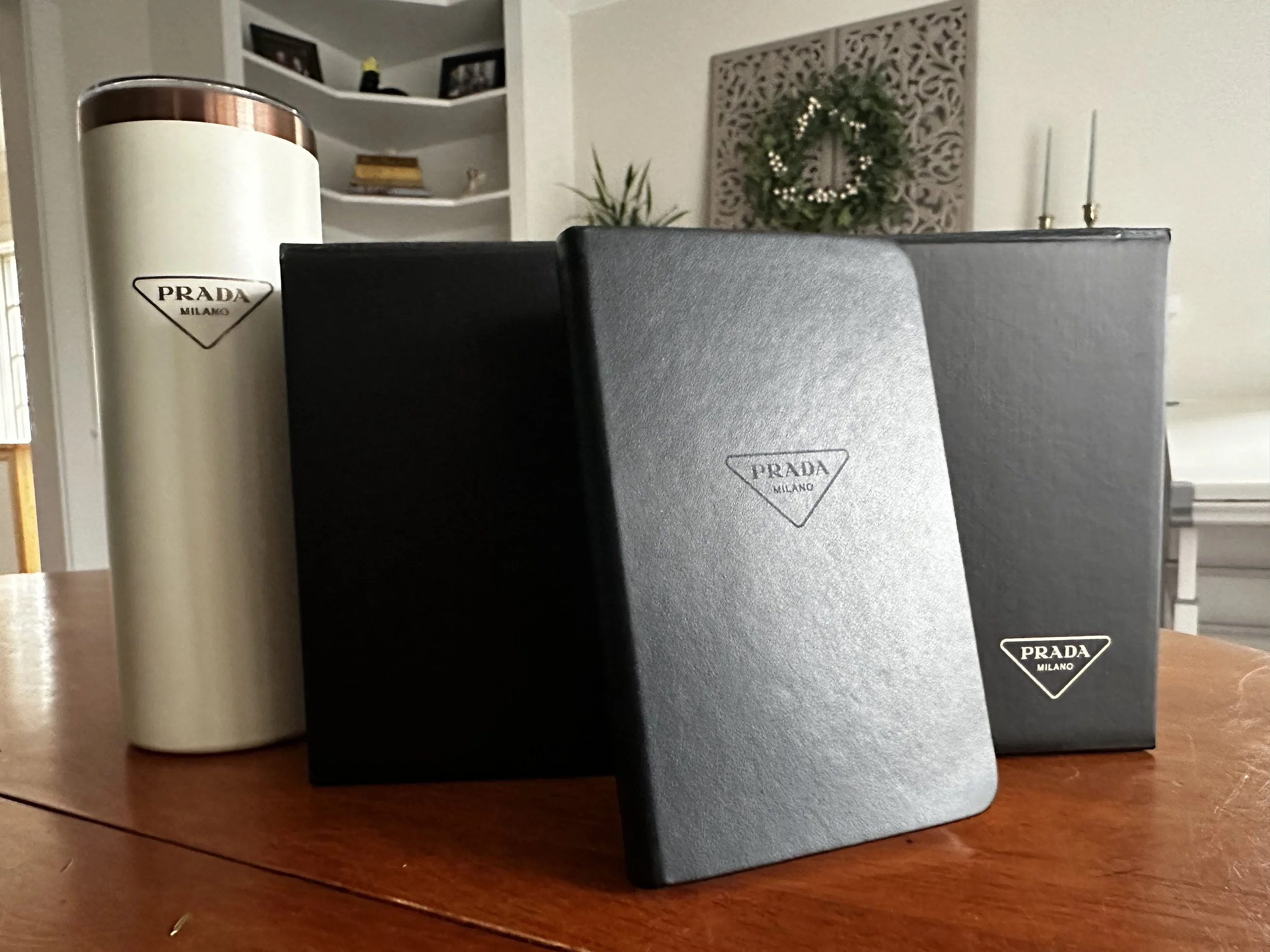 Three luxury Prada Milano boxes in black with silver logo on a wooden table, with a white candle and a black item, possibly a wallet or small bag, also on the table in a room decorated with holiday wreath and candles in the background.