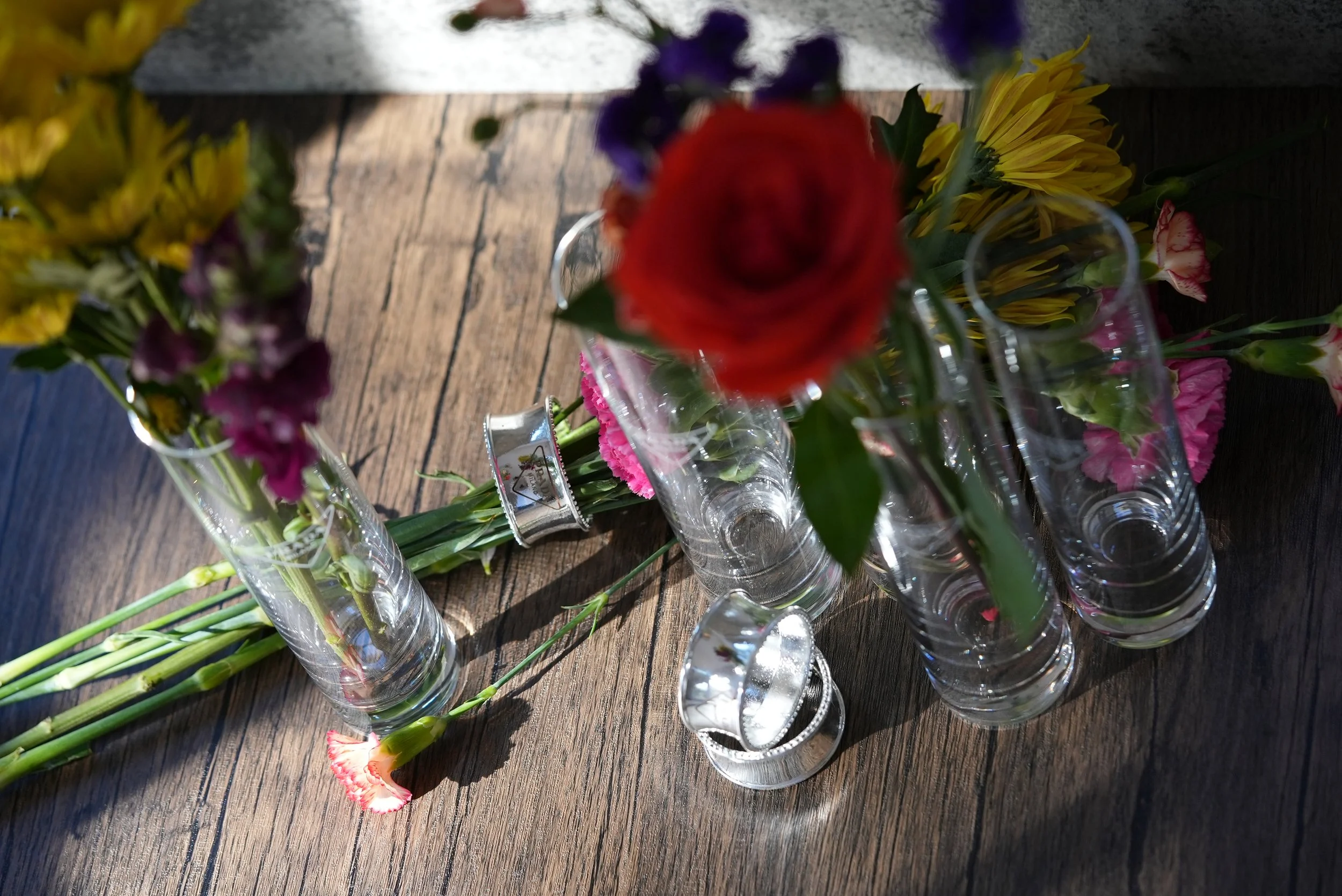 Multiple empty glass vases with flowers placed on a wooden surface, including a red rose, yellow daisies, purple flowers, and pink carnations, with some flowers inside the vases.