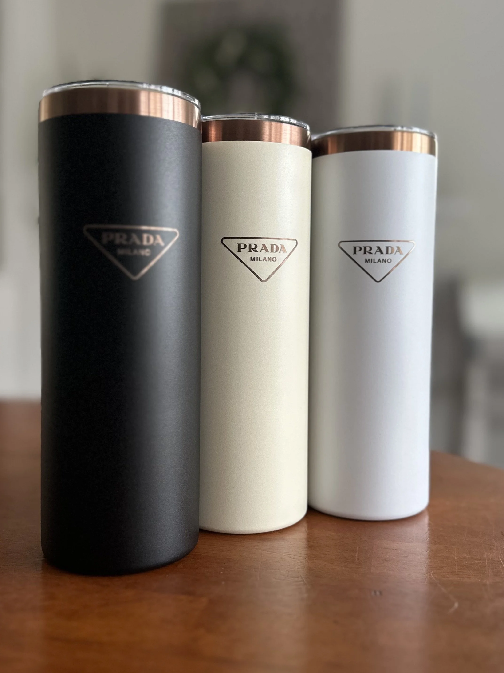 Three Prada Milano perfume bottles with matte finish in black, cream, and white, standing upright on a wooden surface.