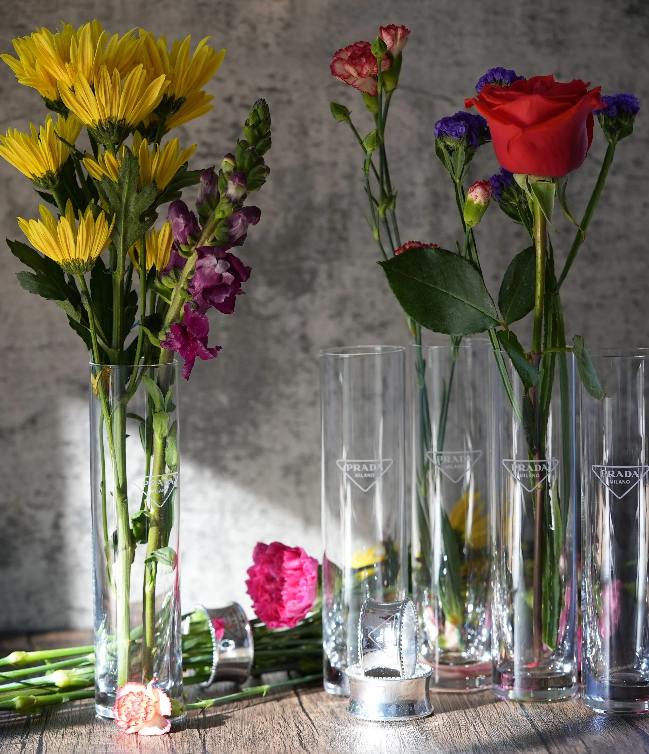 Colorful flowers in clear prada Milano glasses on a wooden surface, grey background, some flowers fallen on the surface.