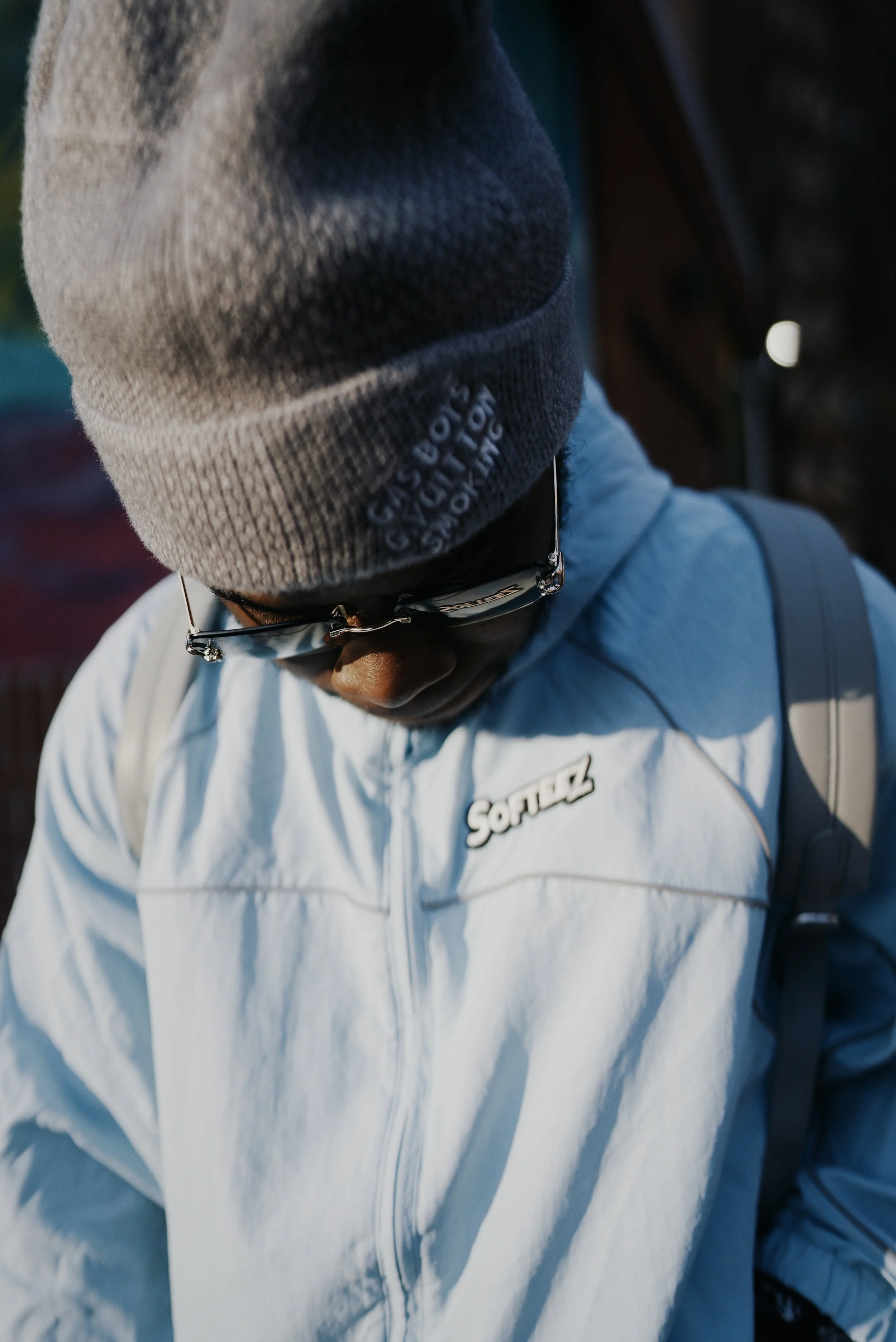 A person wearing a gray knit beanie with black lettering, glasses, and a light blue and white sports jacket.