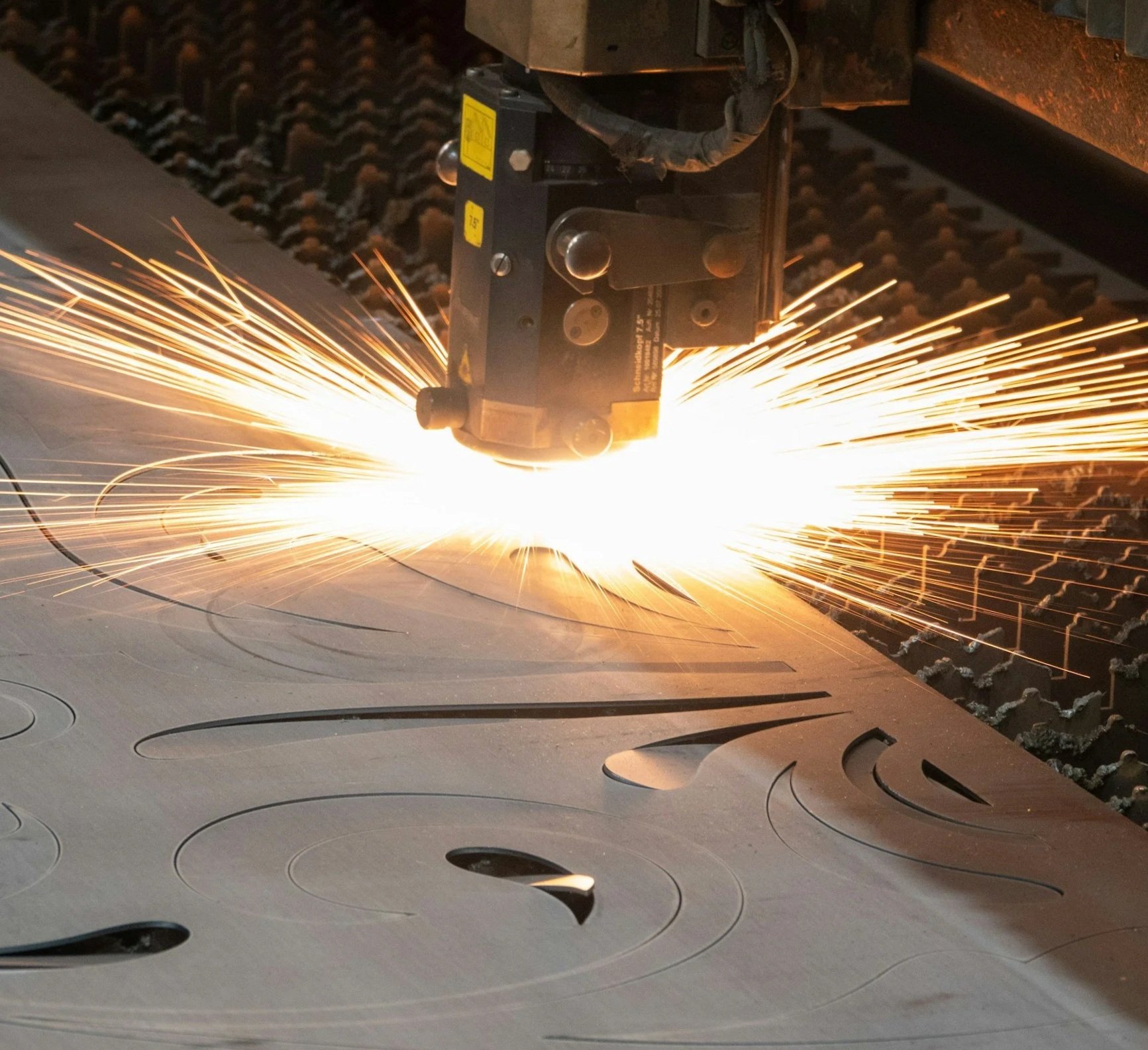 Close-up of a CNC laser cutter machine cutting intricate patterns into metal sheets, with sparks flying from the cutting process.