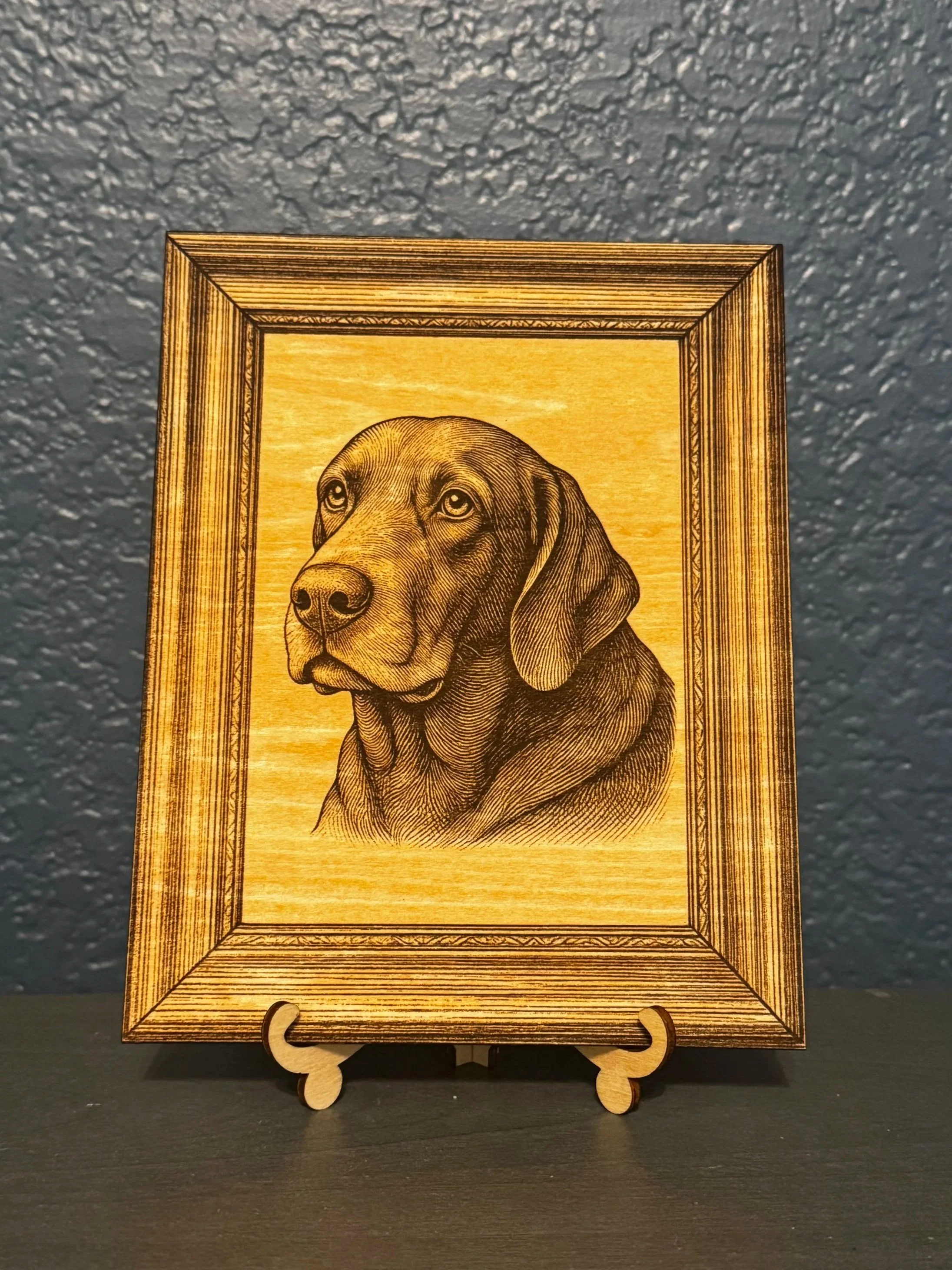 Wooden framed artwork of a Labrador Retriever dog portrait on a display stand, set against a textured gray wall.